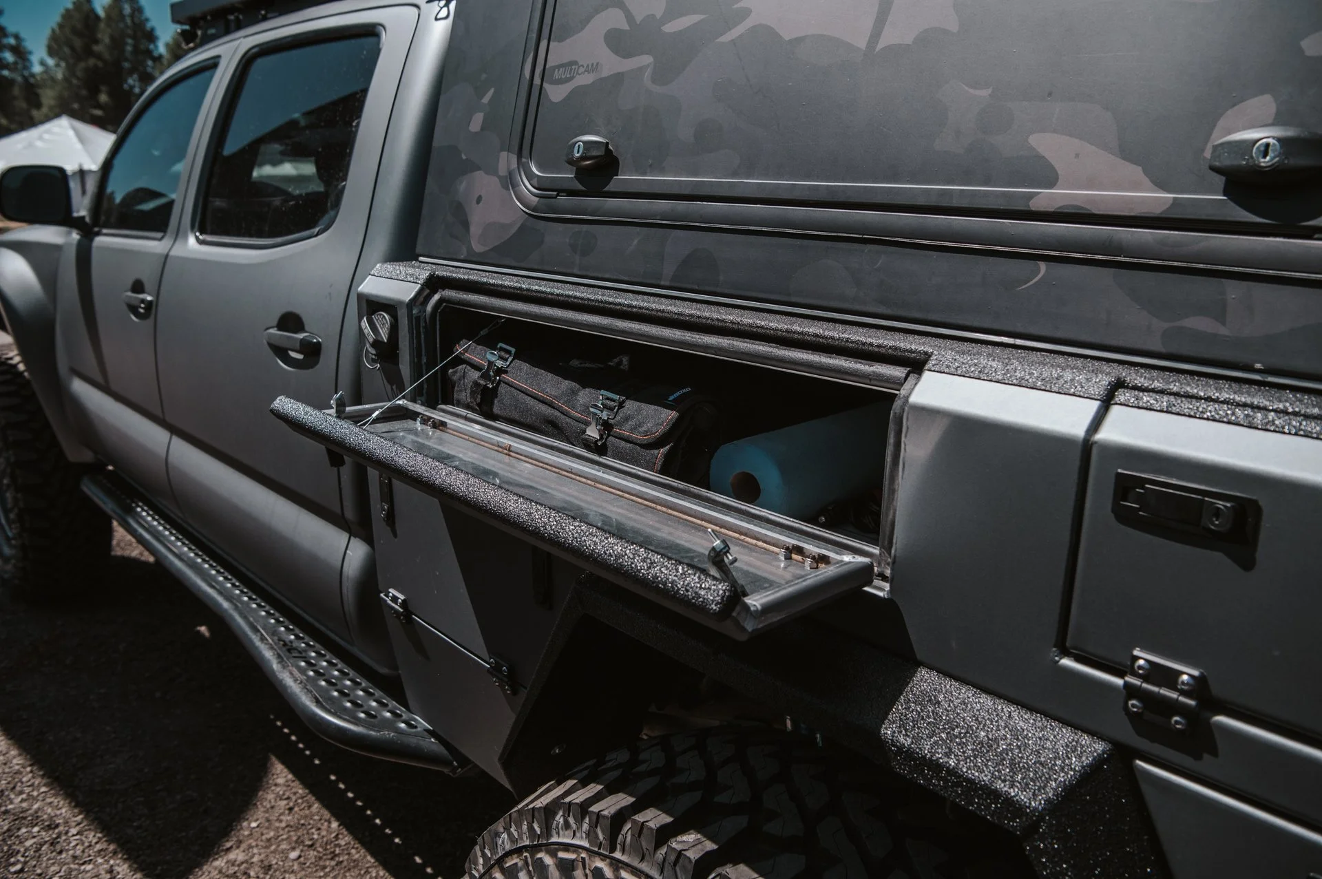 The side of a gray off-road vehicle with an open storage compartment revealing a black bag and a blue rolled-up mat inside.