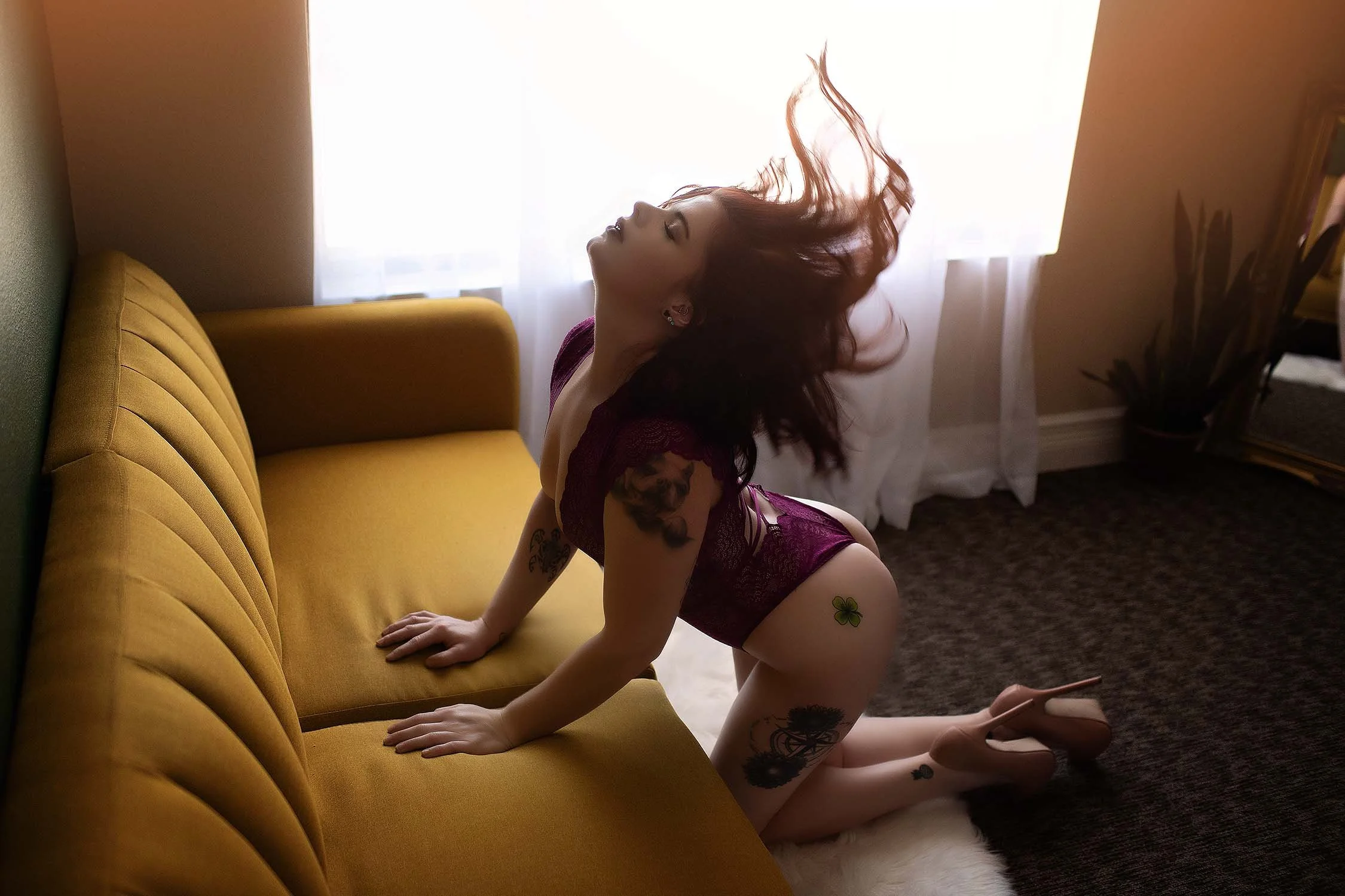 A woman with red hair, tattoos, and wearing purple lingerie, kneeling on a white rug with her hands on a mustard yellow couch, in front of a window with white curtains.