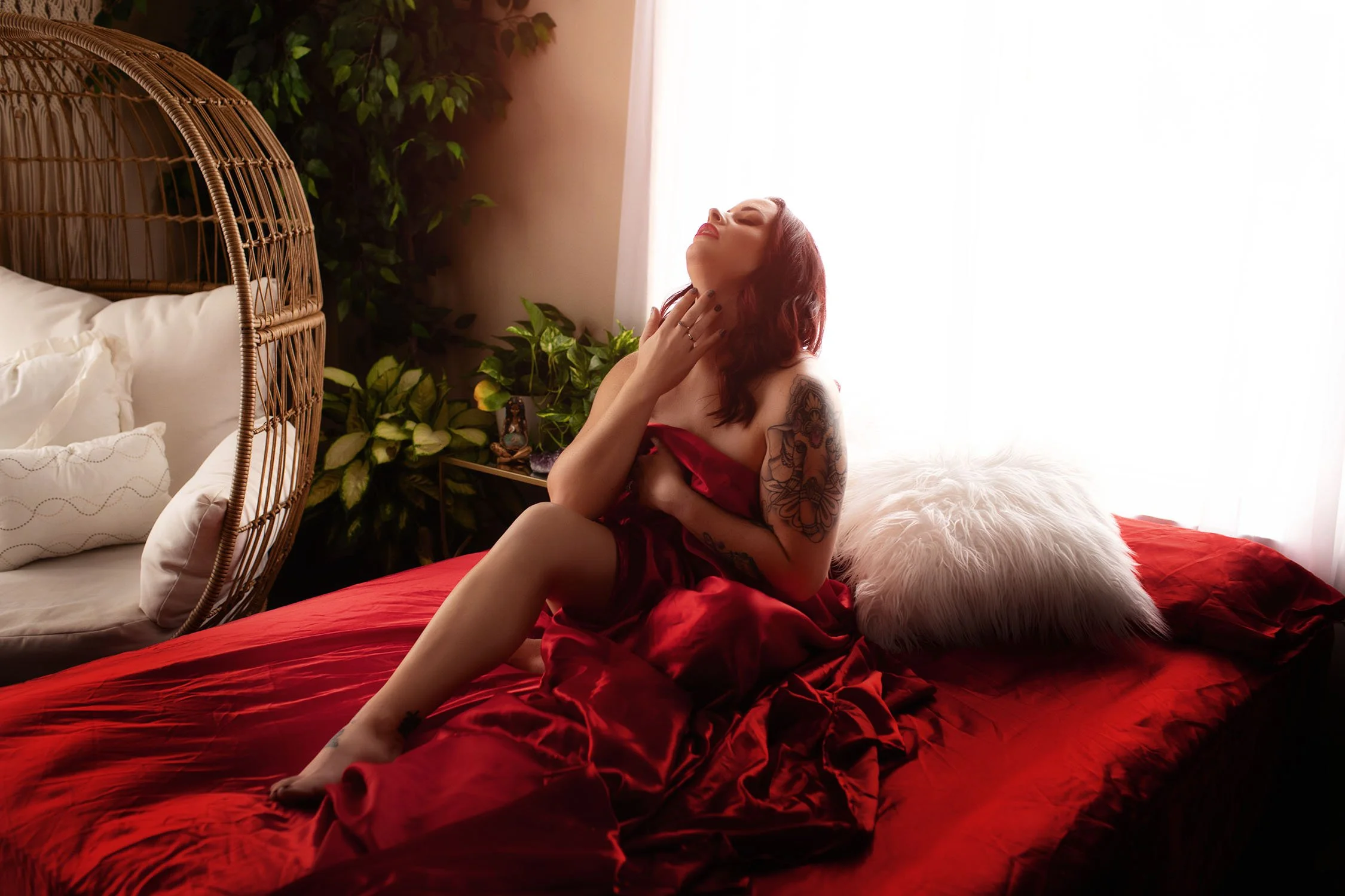 Woman in red satin dress sitting on a bed, with her head tilted back, eyes closed, and hand touching her neck, in a softly lit bedroom with a wicker hanging chair, plants, and a white fluffy pillow.
