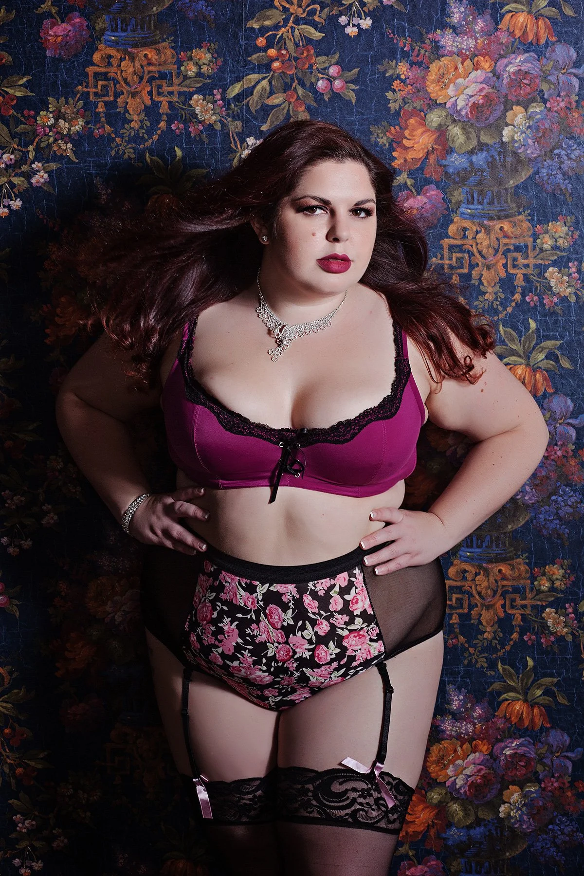 A woman with dark brown hair lying on a floral patterned background, wearing purple and black lingerie, a floral garter belt, and stockings, accessorized with jewelry.