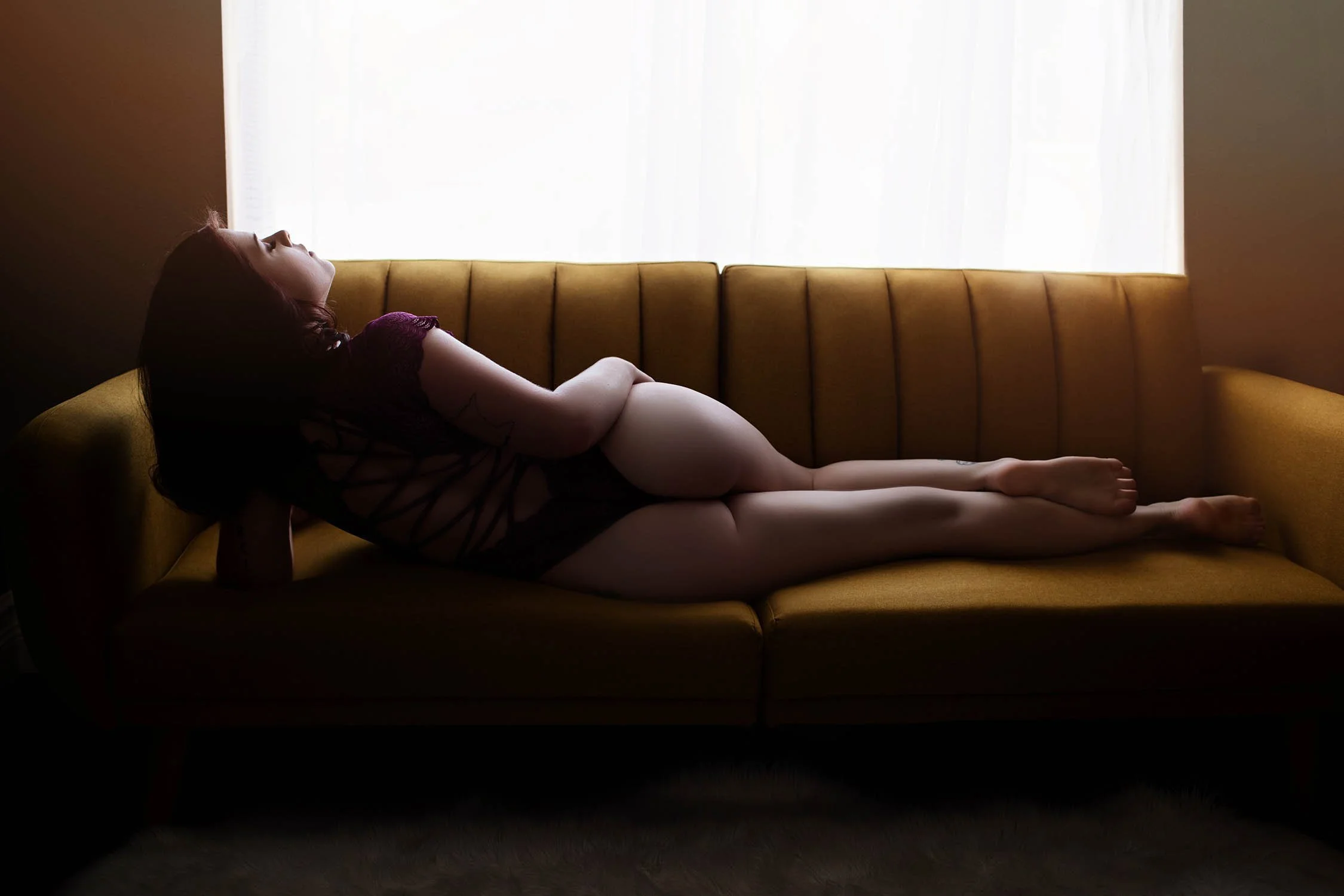 A woman with dark hair lies on a mustard yellow sofa, with her head tilted back and eyes closed, near a bright window with sheer curtains.