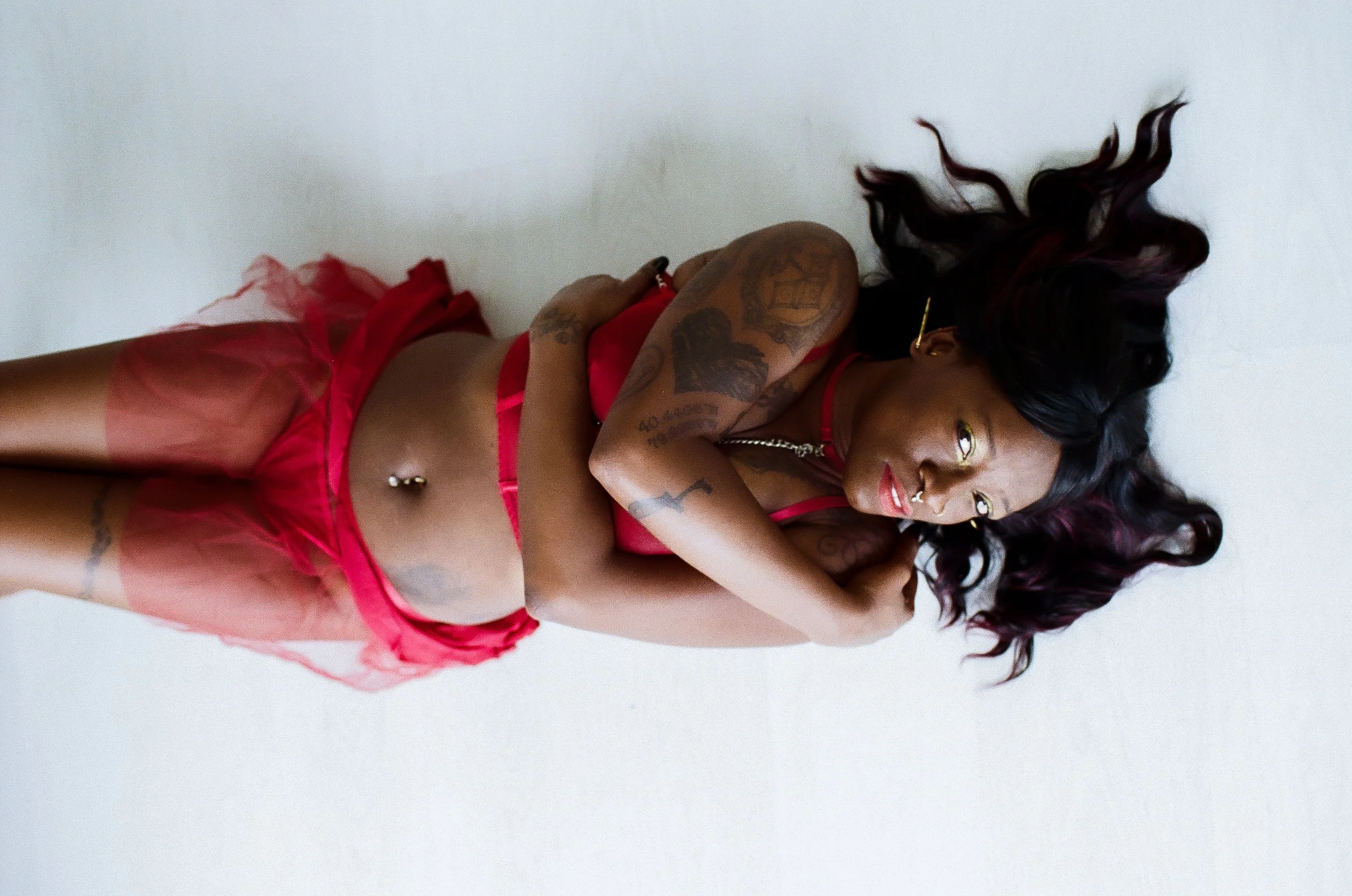 A woman lying on a white surface, wearing red lingerie and sheer pink skirt, with long, dark, wavy hair spread around her head.