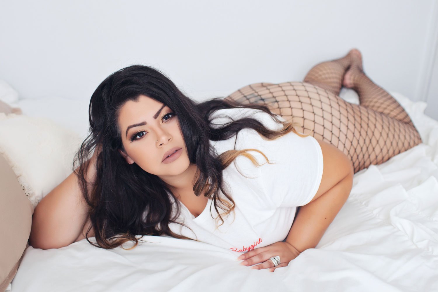 A woman with dark hair, wearing fishnet stockings and a white t-shirt, lying on her side on a bed.