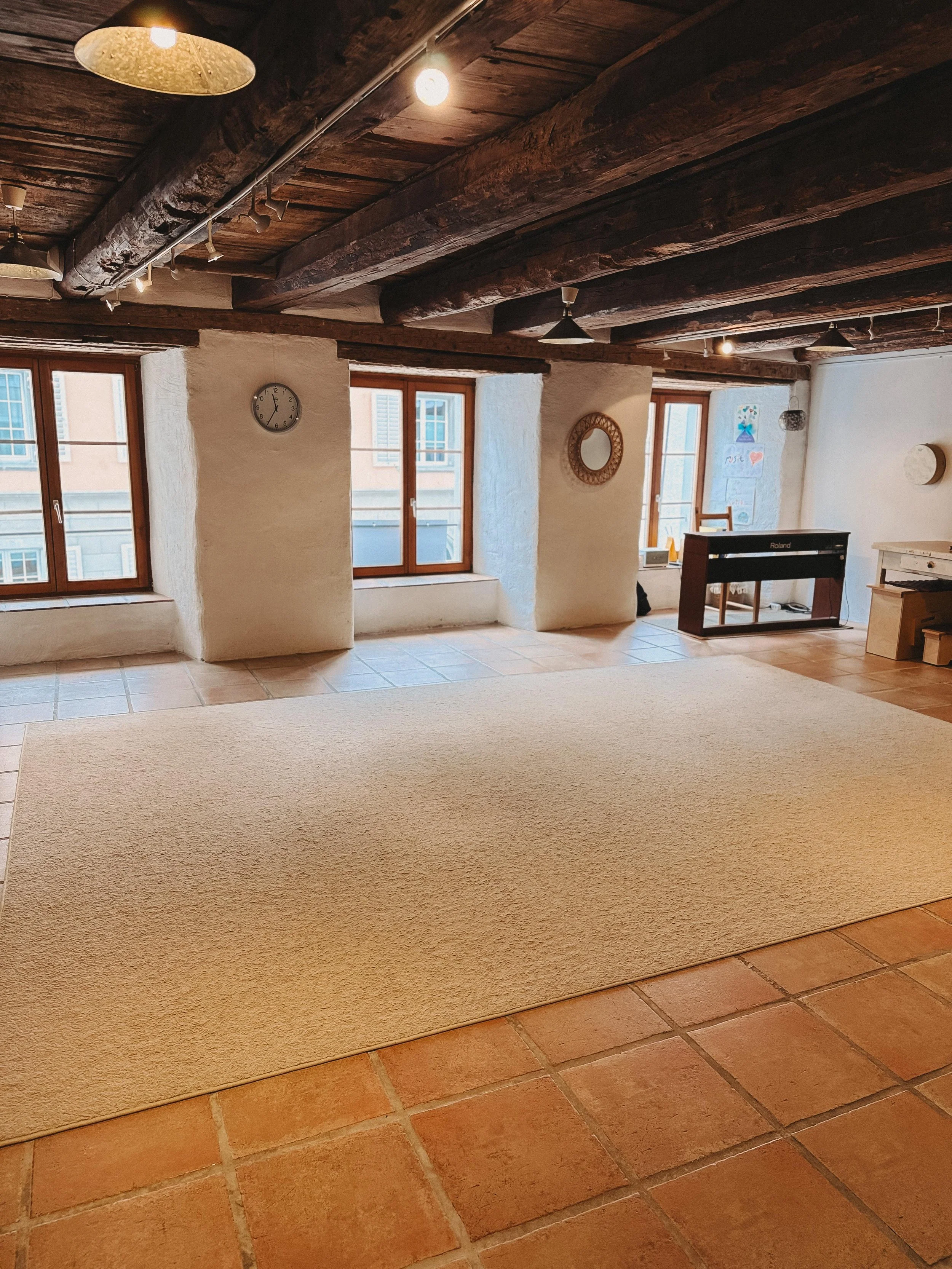 Innenraum eines rustikalen Zimmers mit Holzbalken an der Decke, mehreren Fenstern, einem Wanduhren, einem Spiegel und einem Klavier, mit Fliesenboden und einem Teppich.