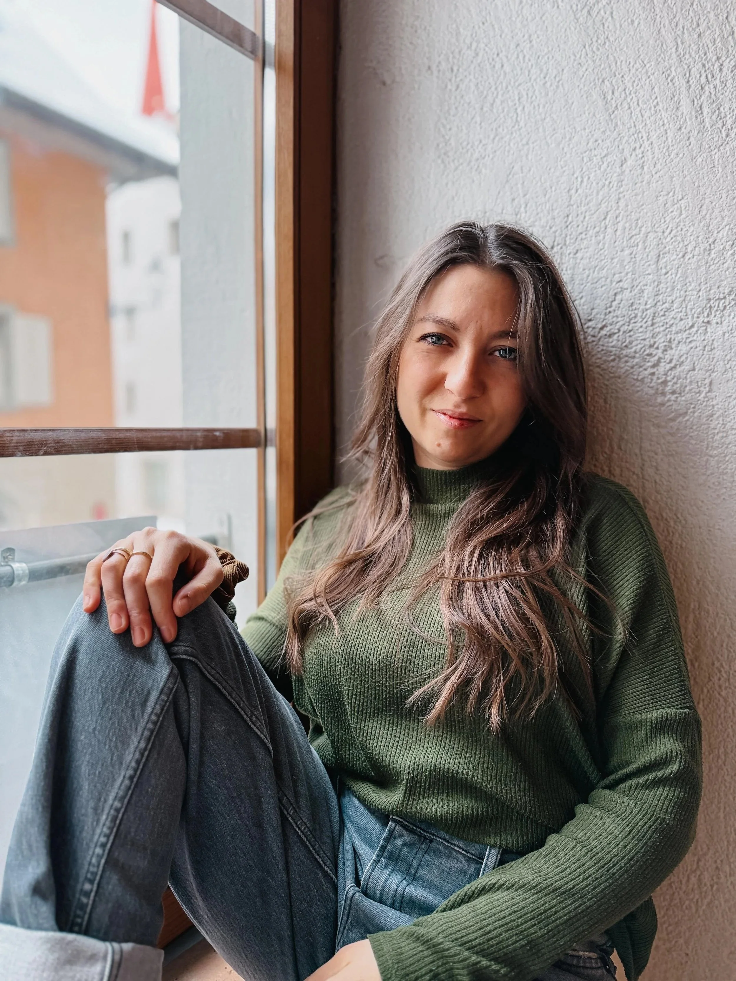 Junge Frau mit braunen Haaren und blauen Augen sitzt an einem Fenster, trägt ein grünes Pullover und blaue Jeans, lächelt leicht.