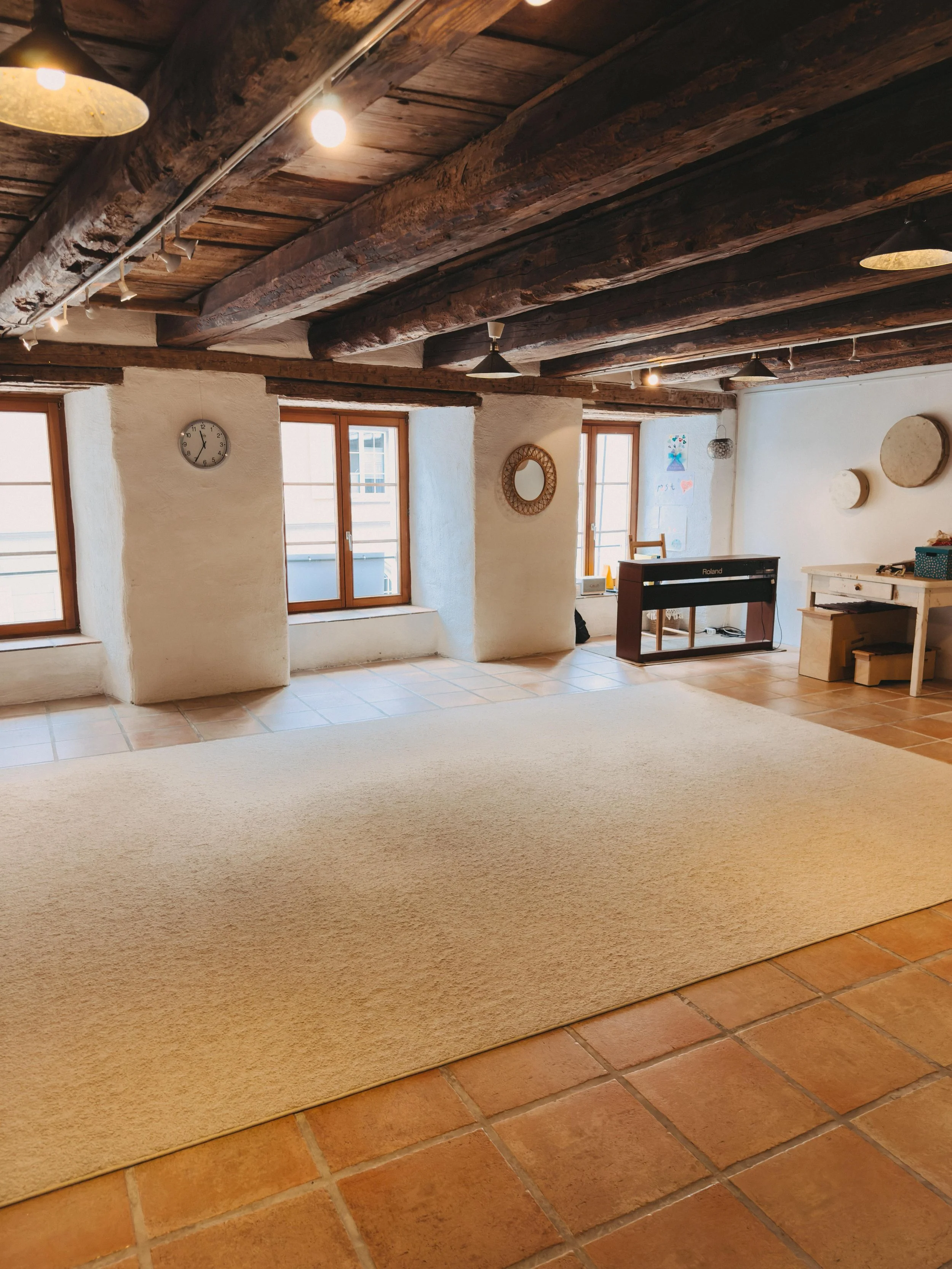 Innenraum mit rustikaler Decke und großen Fenstern, die Tageslicht hereinlassen. Es gibt einen Klavier, einen Schreibtisch, einige Dekorationsstücke an der Wand und einen Teppich auf Fliesenboden.