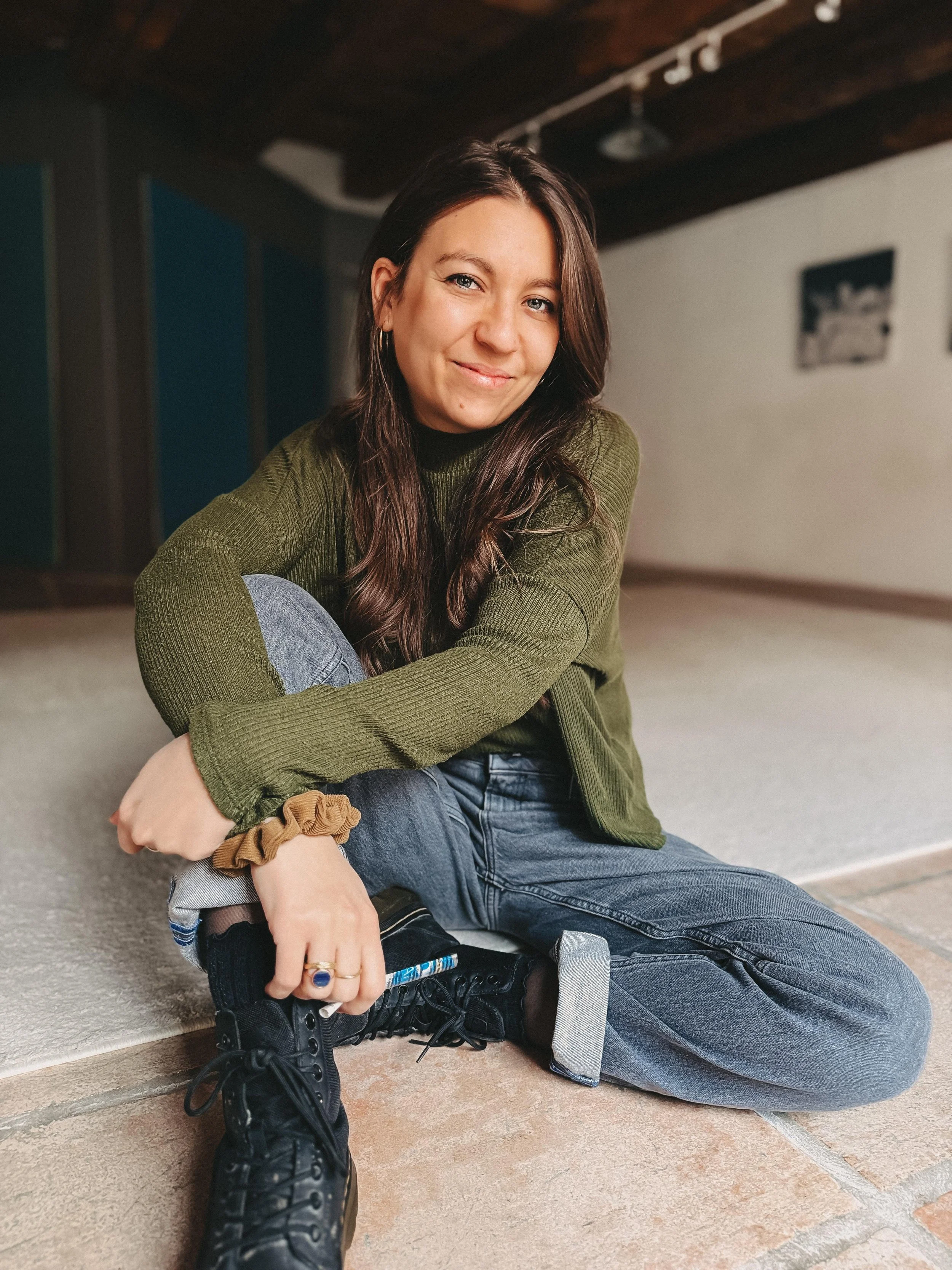 Junge Frau sitzt auf dem Boden, trägt eine grüne Strickjacke, Jeans, schwarze Schuhe, hält einen Marker in der Hand und lächelt.