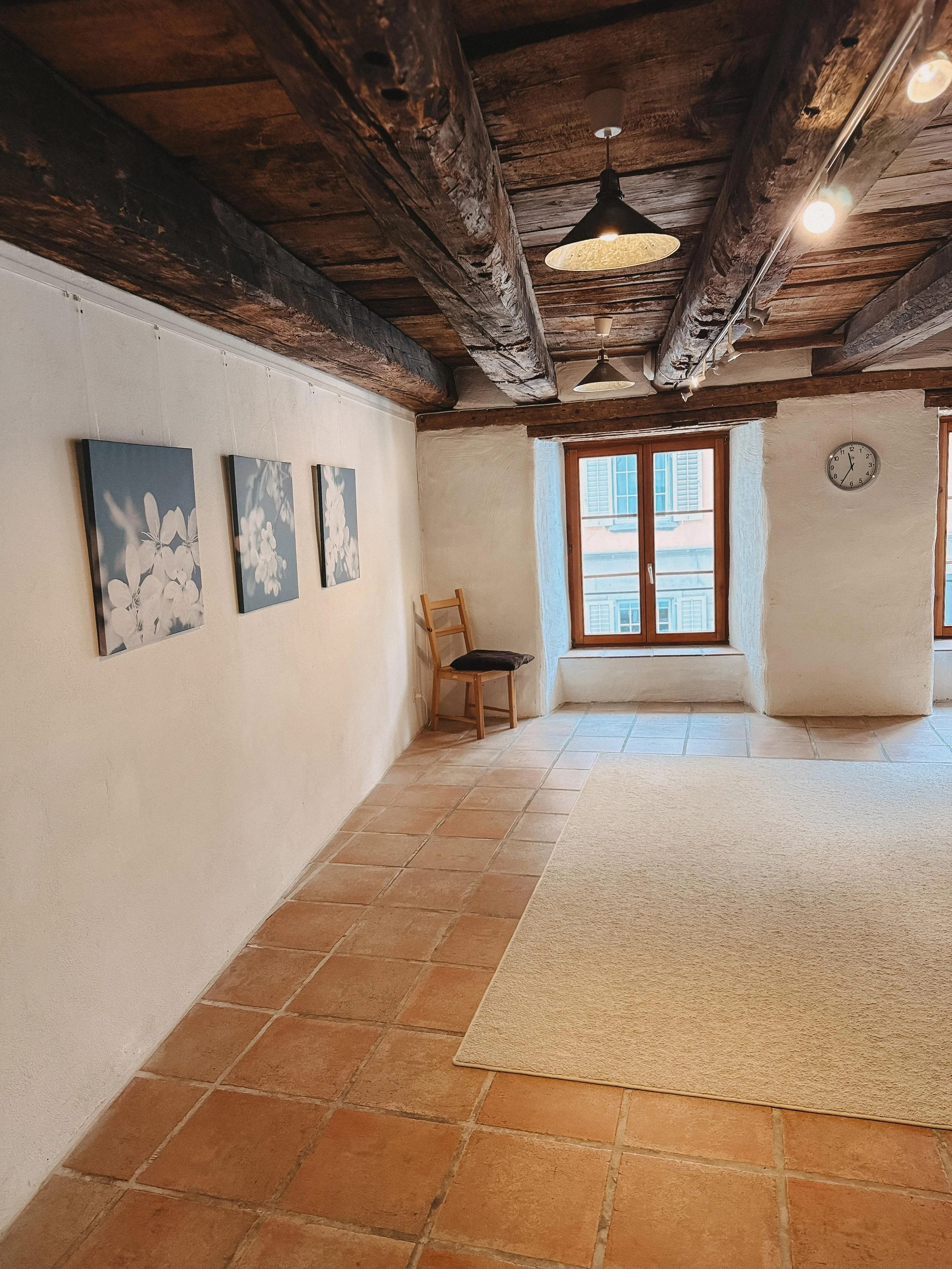 Ein Raum mit weißen Wänden, einem Holzfenster, Terrakottabodenfliesen, einem kleinen Holzstuhl, drei Wandbilder mit Blumenmotiven, einer Deckenleuchte und einer Uhr an der Wand.