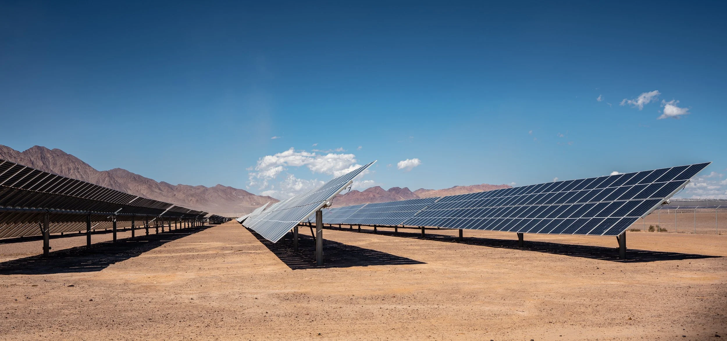 Solar panels in a desert landscape with mountains in the background under a blue sky.
