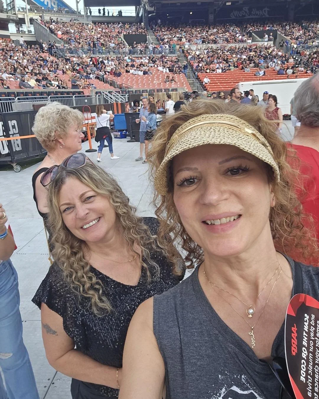 A man with curly black hair, glasses, and a beard, wearing a dark green sweater, looking to the side with a neutral expression, indoors with a plant and a white wall in the background.