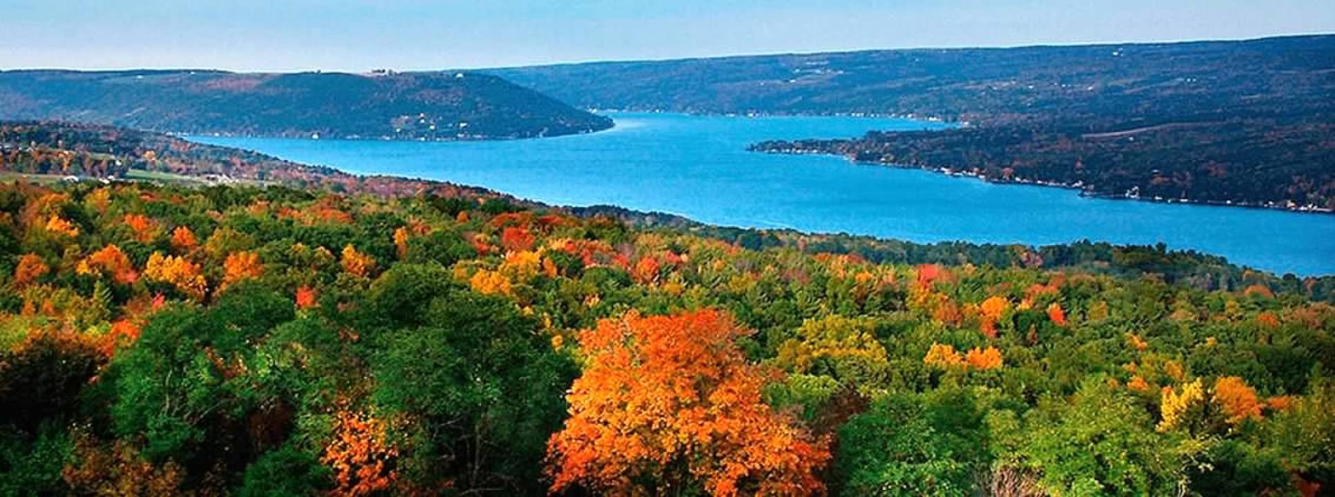 A scenic view of a large lake surrounded by hills and dense forest with colorful autumn foliage in the foreground.