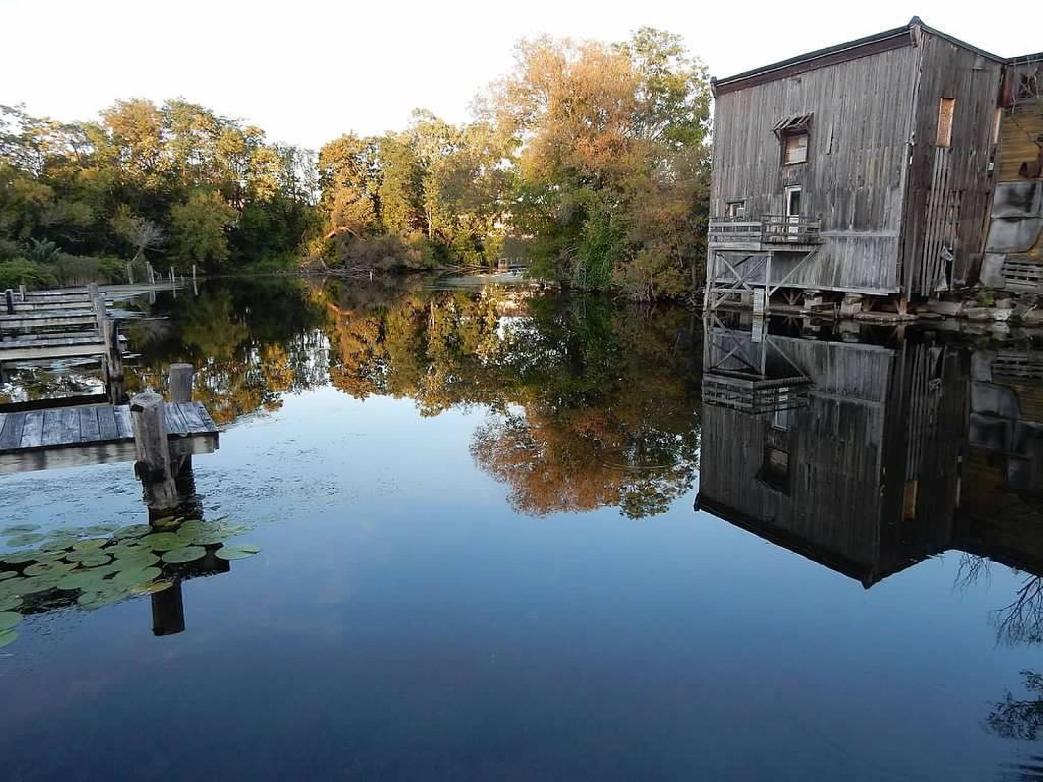 A calm body of water reflects a wooden building and autumn trees with green, yellow, and orange leaves, along with a dock on the left side of the image.