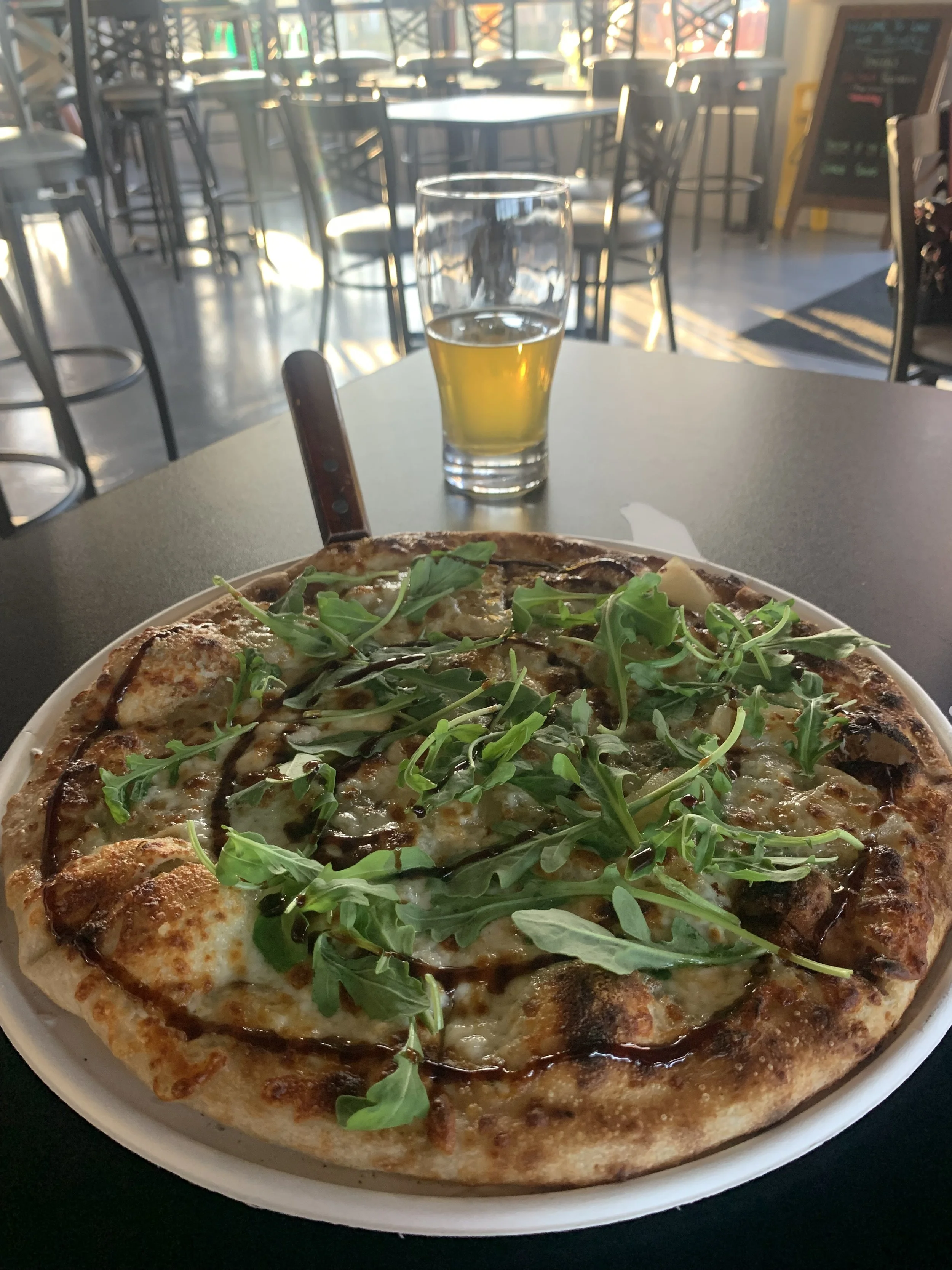 A pizza topped with arugula leaves, cheese, and balsamic glaze on a white plate, with a glass of beer on the table in the background inside a restaurant.