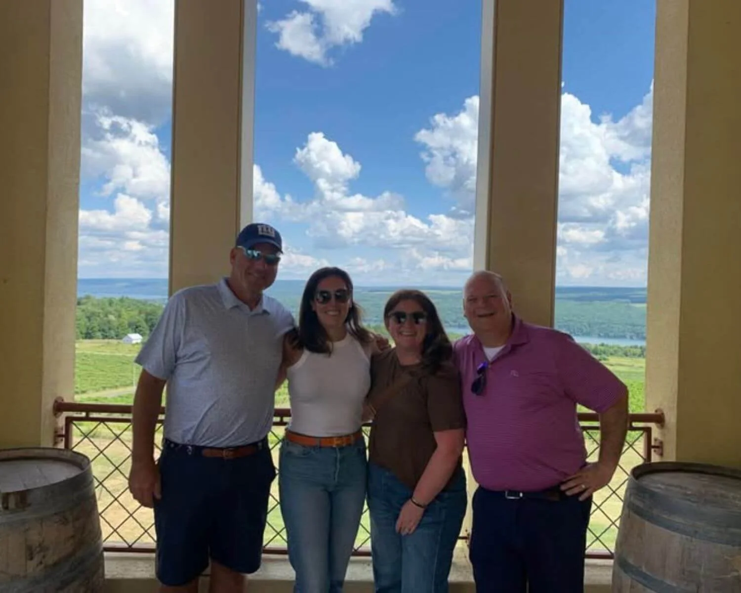Four people standing together on a balcony with scenic countryside and a river in the background, smiling and posing for a photo.