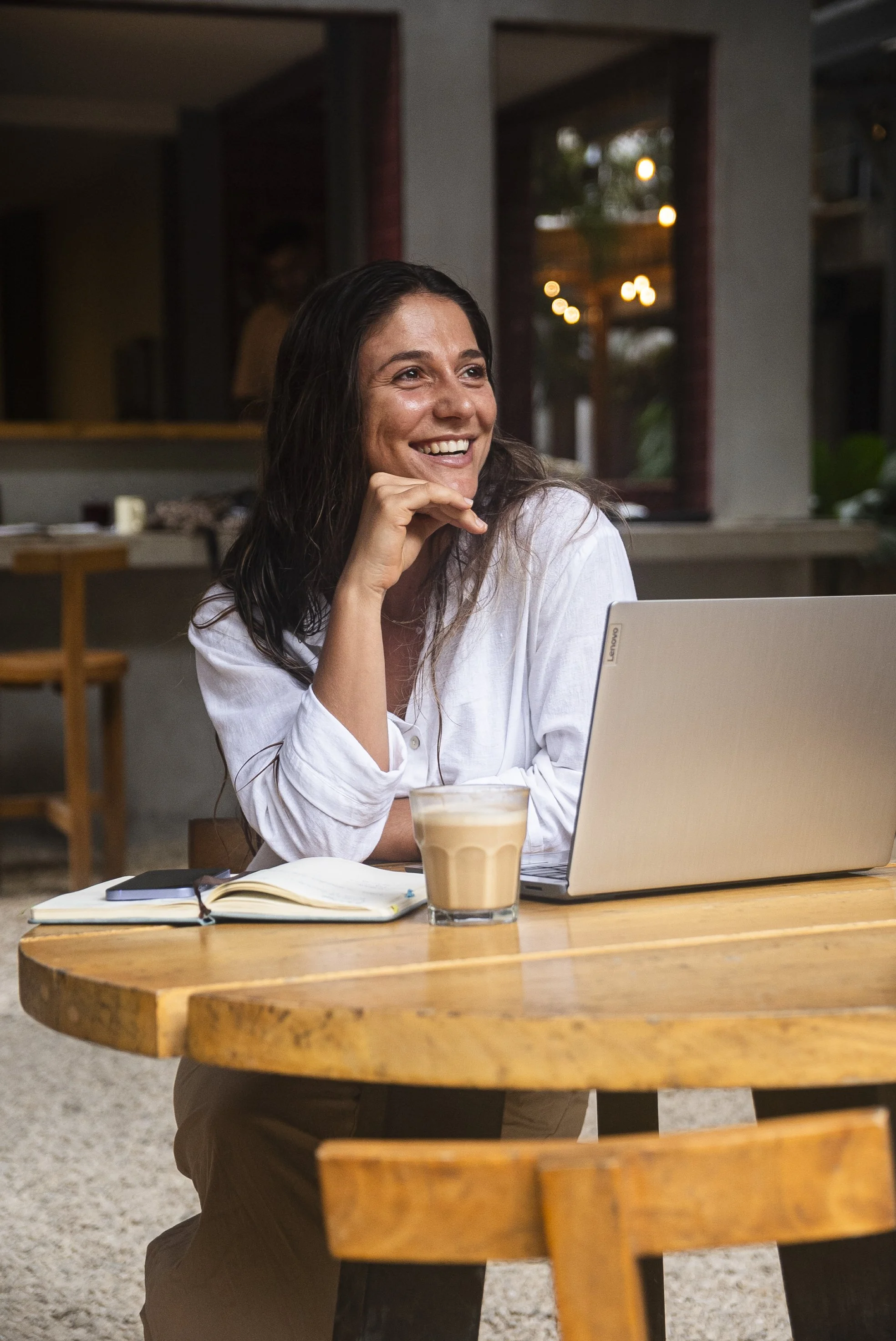 Una mujer sonriente sentado en una mesa de madera con una laptop, una taza de café con leche, un cuaderno y un teléfono en un ambiente acogedor de cafetería. Lista para impartir sus clases en linea.
