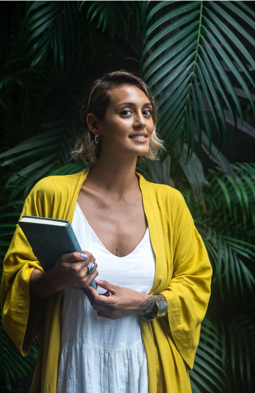 Mujer joven con cabello corto, luciendo una blusa blanca y un cardigan amarillo, sosteniendo un cuaderno y sonriendo, con fondo de hojas verdes y plantas tropicales.