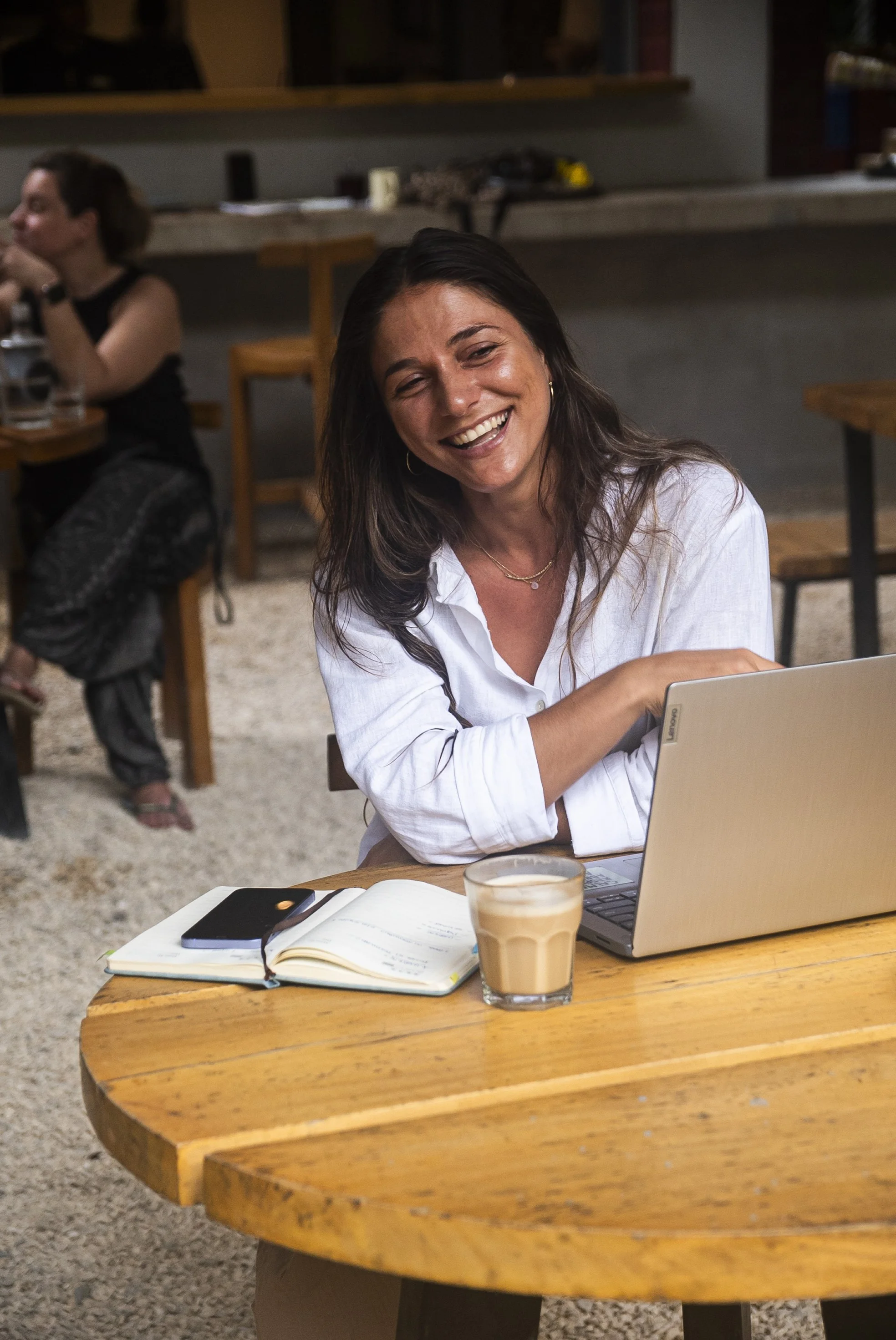 Mujer sonriendo sentada en una mesa de madera en una cafetería, con una laptop, una taza de café con leche y un cuaderno con notas.