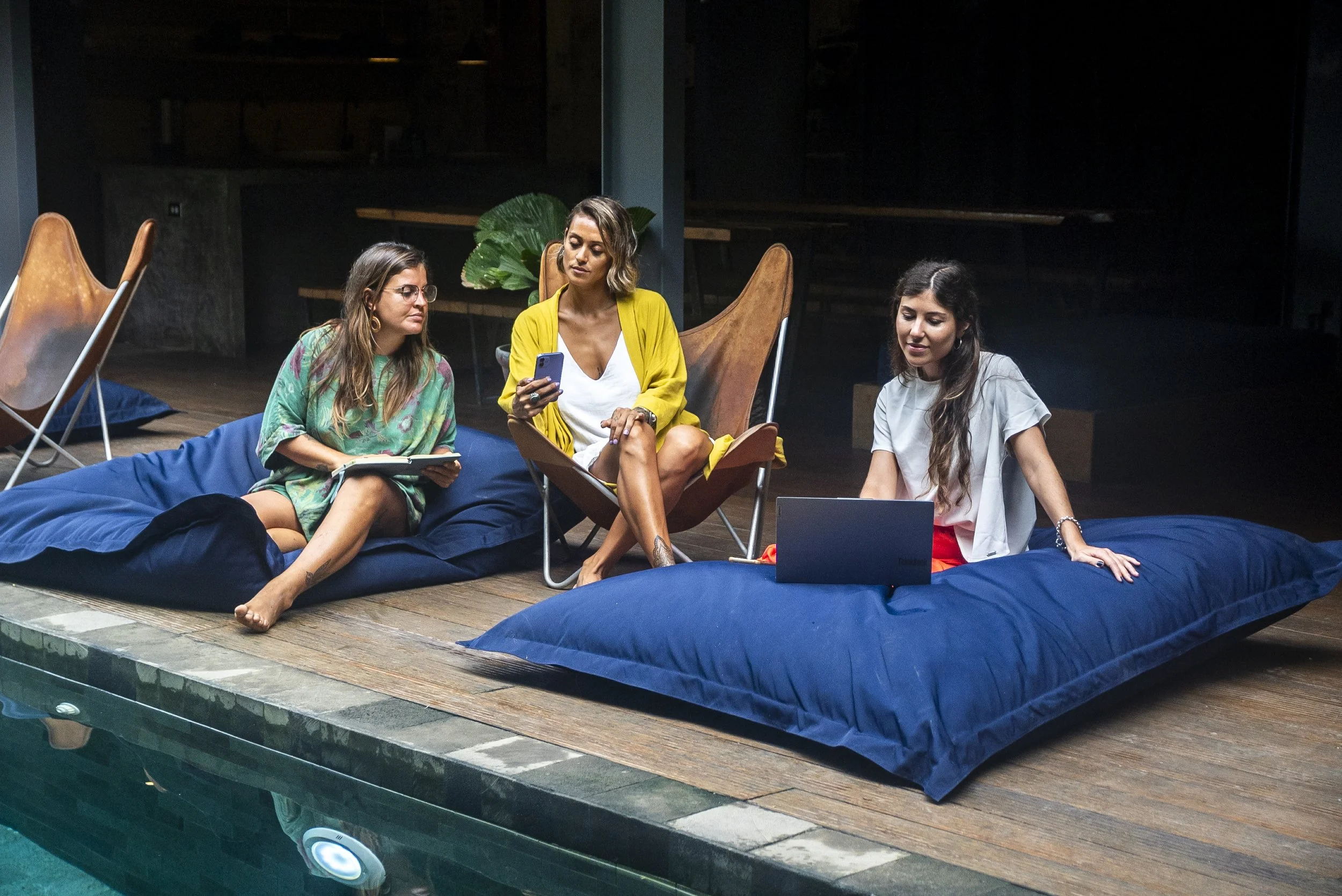 Tres mujeres en un espacio interior cercano a una piscina, una usan un portátil, otra usa un teléfono móvil y la tercera lee un cuaderno. Están sentadas sobre cojines grandes y decoraciones modernas y naturales en el fondo.