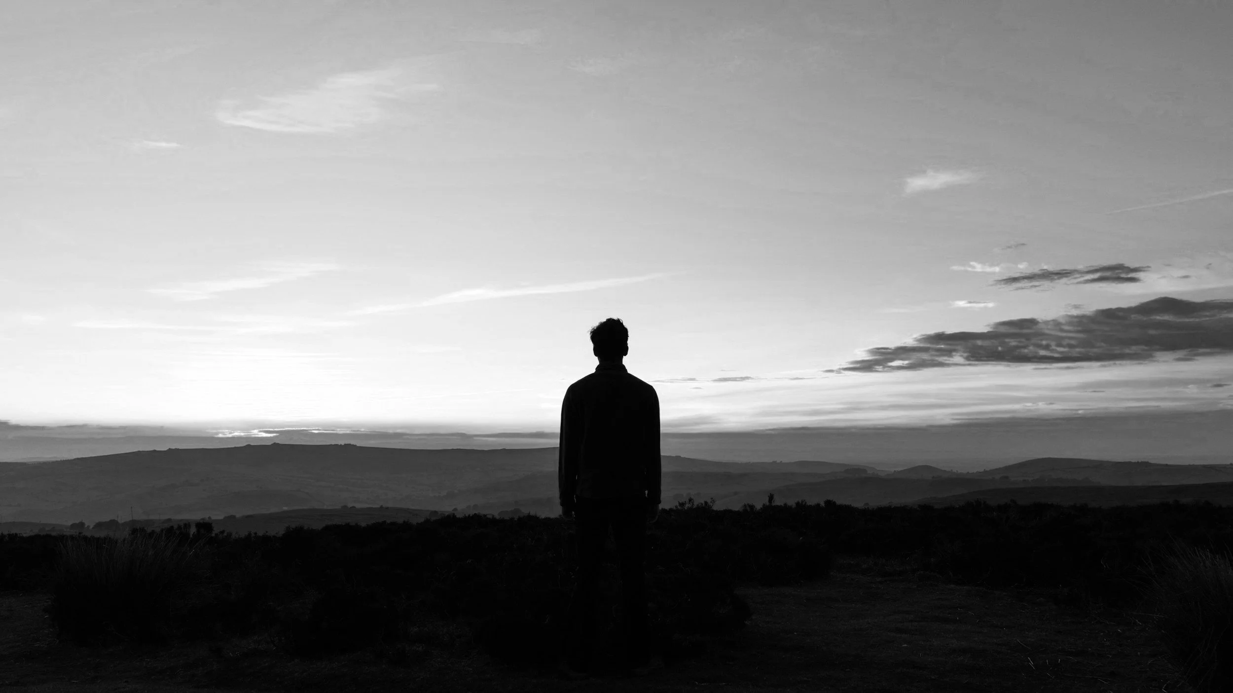 Persona de espaldas observando un paisaje al atardecer en un campo abierto con colinas y cielo con nubes.