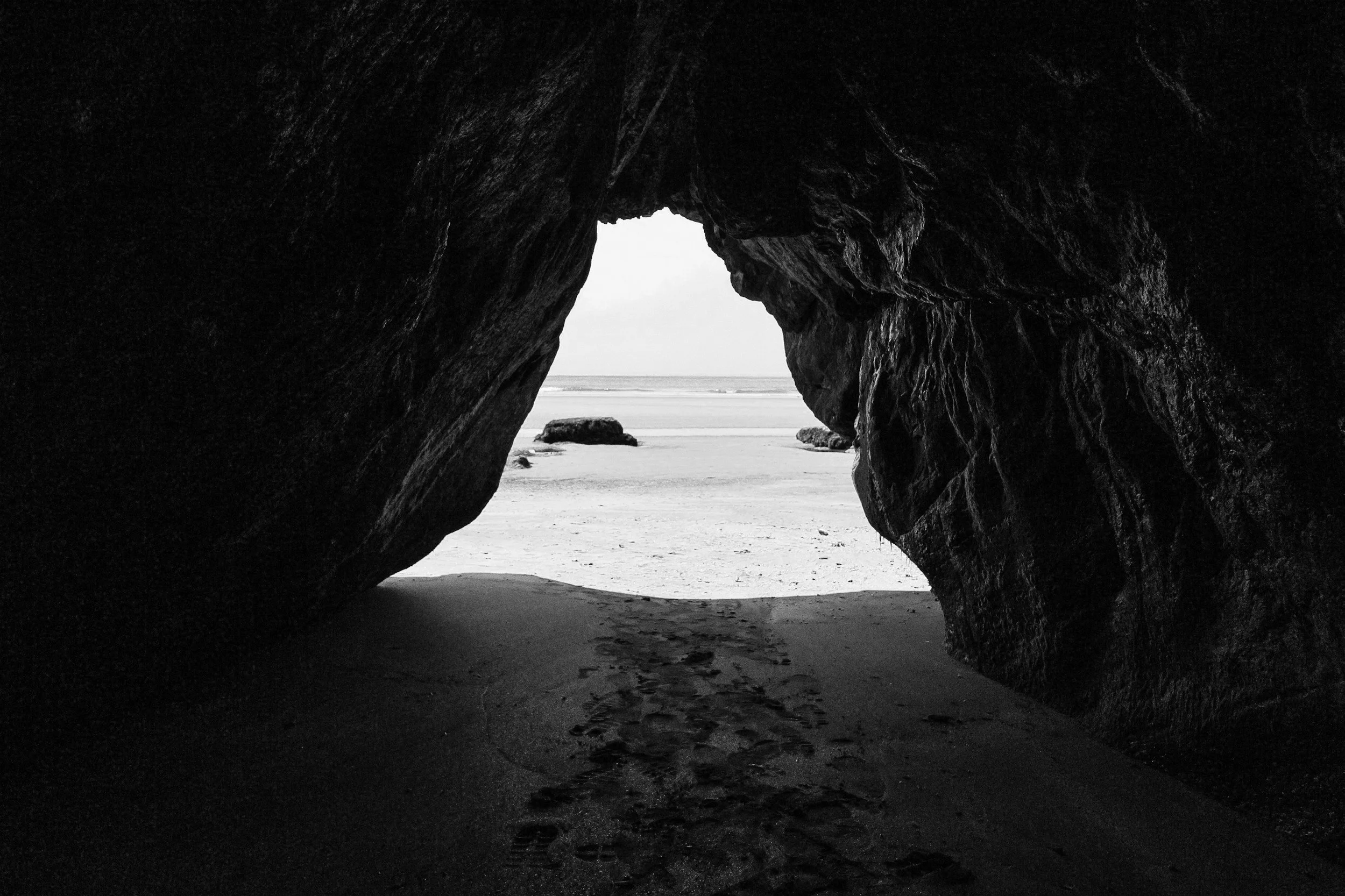 Vista desde una cueva en la playa, con rastro de huellas en la arena y rocas en el fondo, en blanco y negro.