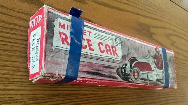 Vintage miniature toy race car packaged in a worn cardboard box labeled 'Mini Race Car' with an illustration of the car, secured with two blue rubber bands.