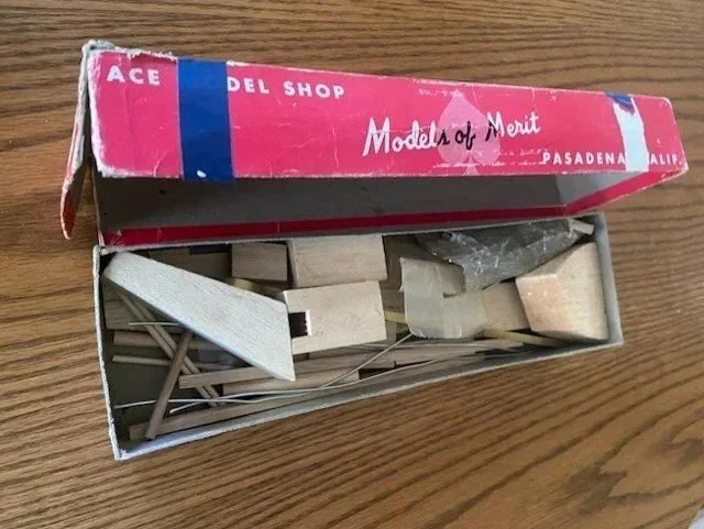 Open vintage red and blue box labeled 'Models of Merit' containing wooden model parts on a wooden surface.