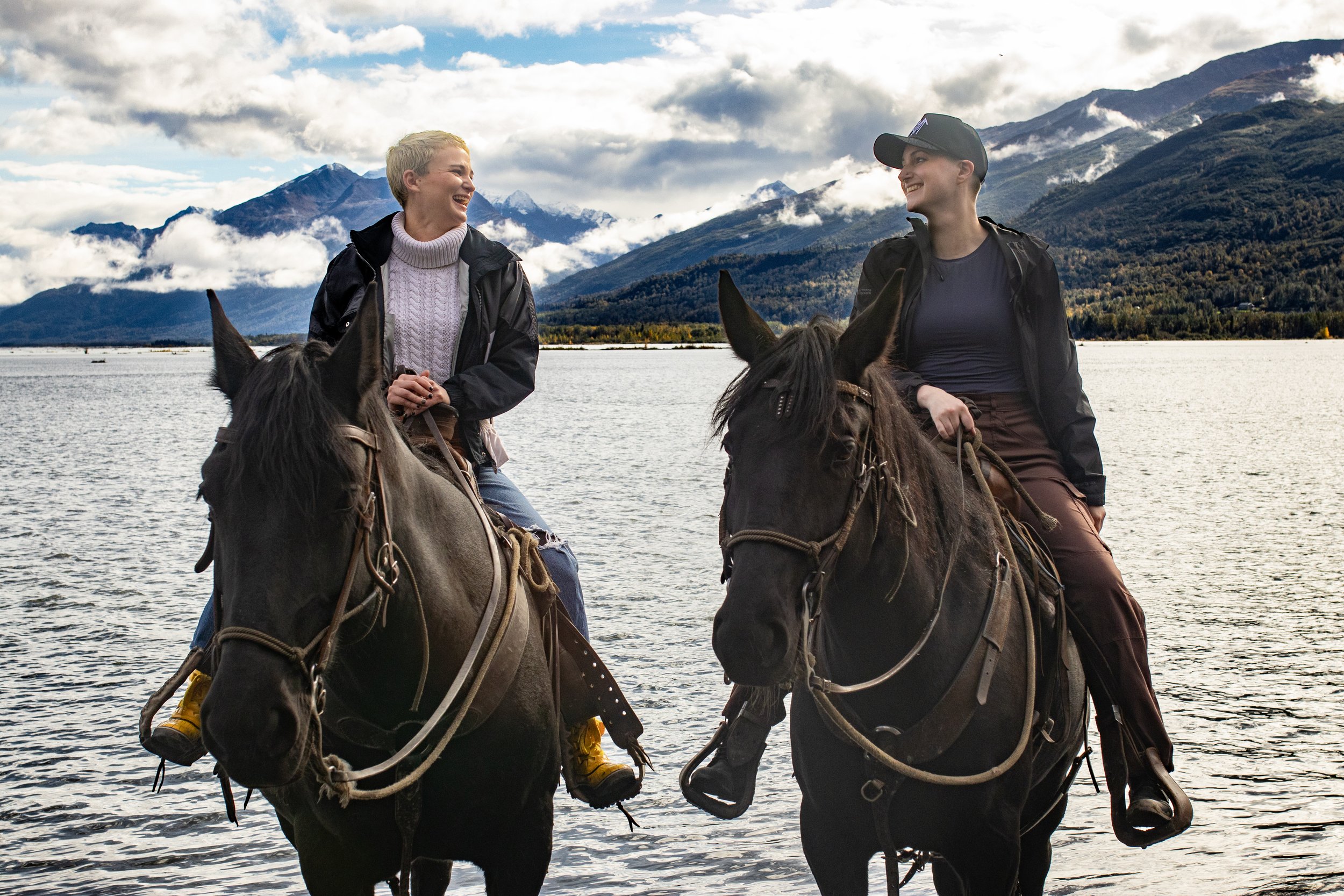 Two women riding horses are standing in a body of water with mountains and clouds in the background, smiling and engaging in conversation in Palmer, Alaska