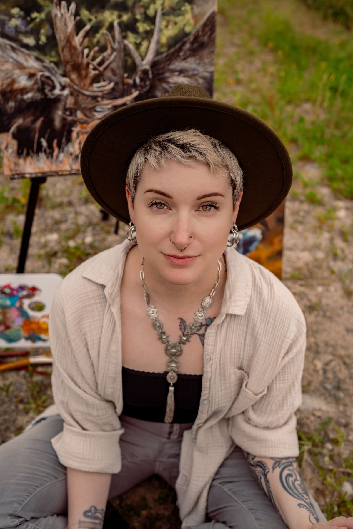 A young woman with short blonde hair, wearing a large brown hat, a nose piercing, and multiple tattoos, sits outdoors with a colorful painting of a moose in the background.