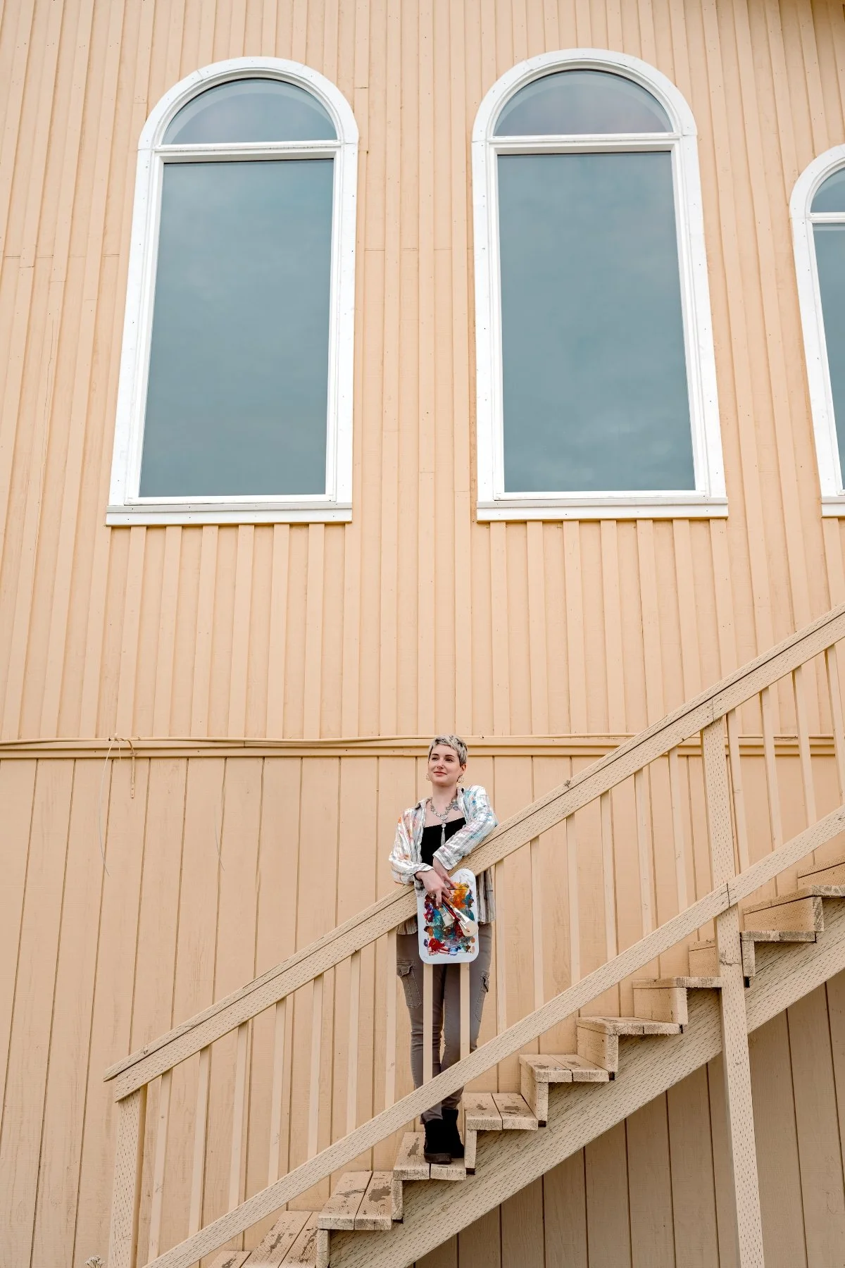 A woman standing on wooden stairs in front of a yellow wooden building with three tall, arched windows.