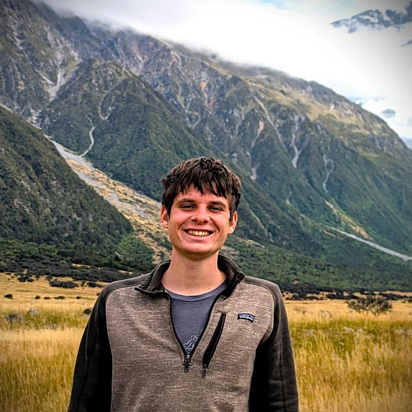 A smiling young man with short brown hair standing in a grassy field with mountains in the background.