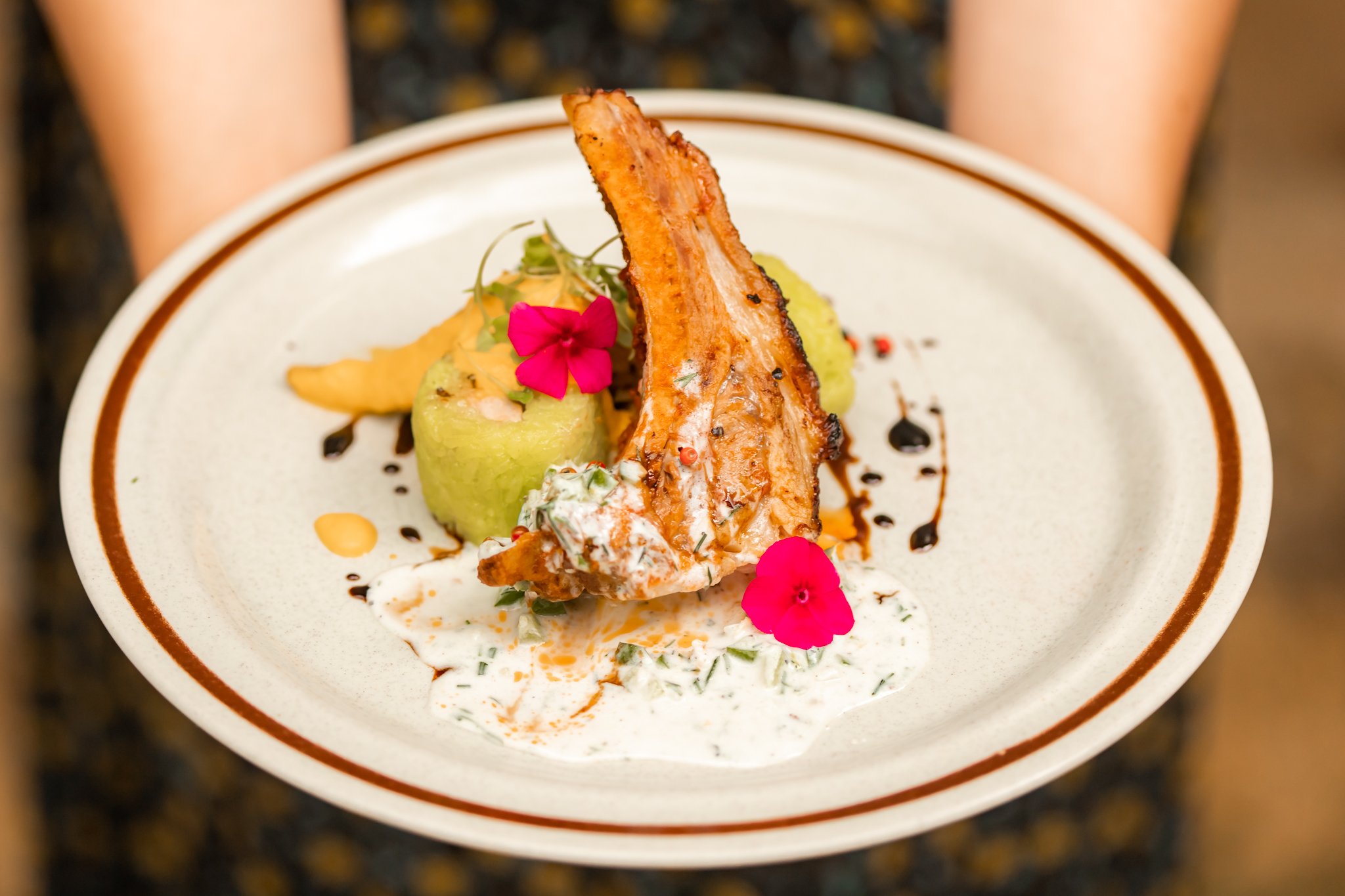 Fine dining plate captured by Maksym Brukhno — grilled salmon garnished with edible flowers, herb sauce, and a carved melon ball. Styled hospitality food photography on a white plate with brown rim.
