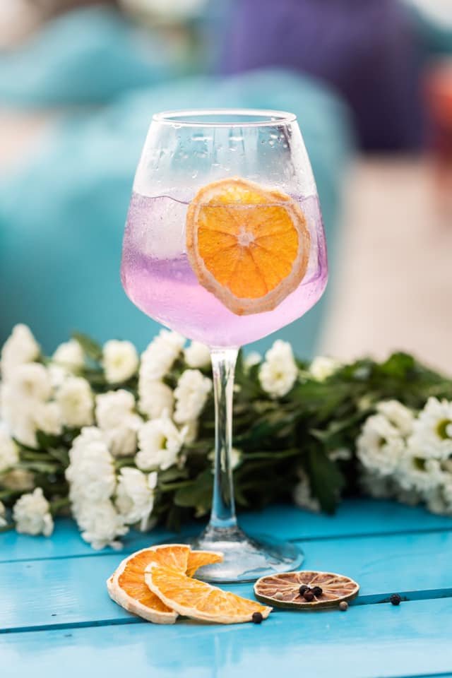 Vibrant summer cocktail with pink liquid, orange slice, and dried citrus garnish — styled on a blue tabletop with florals. Refreshing horeca beverage photography by Maksym Brukhno.