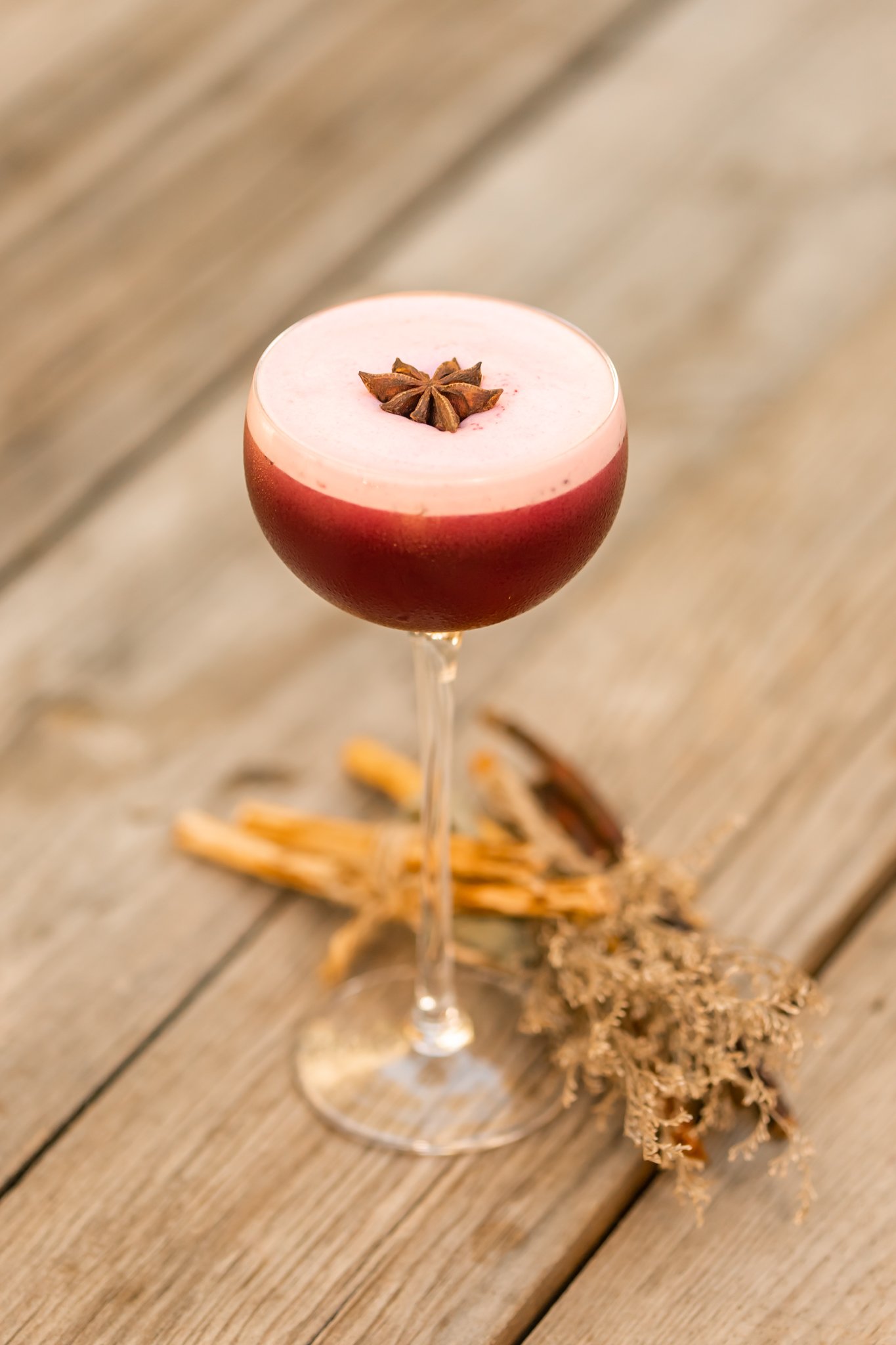 Vibrant pink and red cocktail garnished with star anise, served in a coupe glass on rustic wood, styled with dried botanicals and wood shavings. Photographed by Maksym Brukhno for horeca branding.