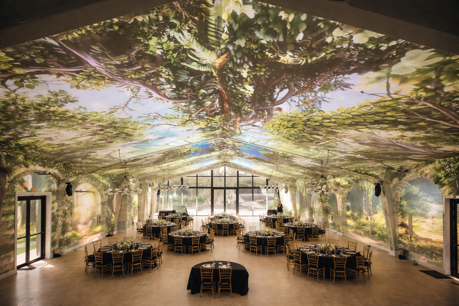 Event hall decorated with walls and ceiling featuring a large-scale nature mural of trees and leaves, set up with round tables draped in black tablecloths, surrounded by gold chairs, and floral centerpieces, with a large glass window at the back letting in natural light.