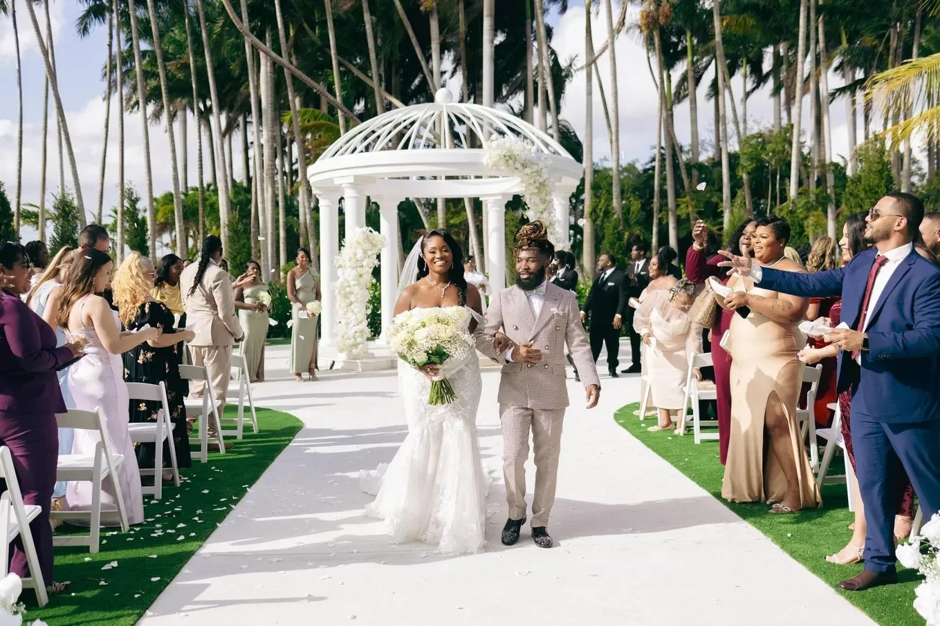 A bride and groom walking down the aisle at an outdoor wedding ceremony, surrounded by guests and palm trees.