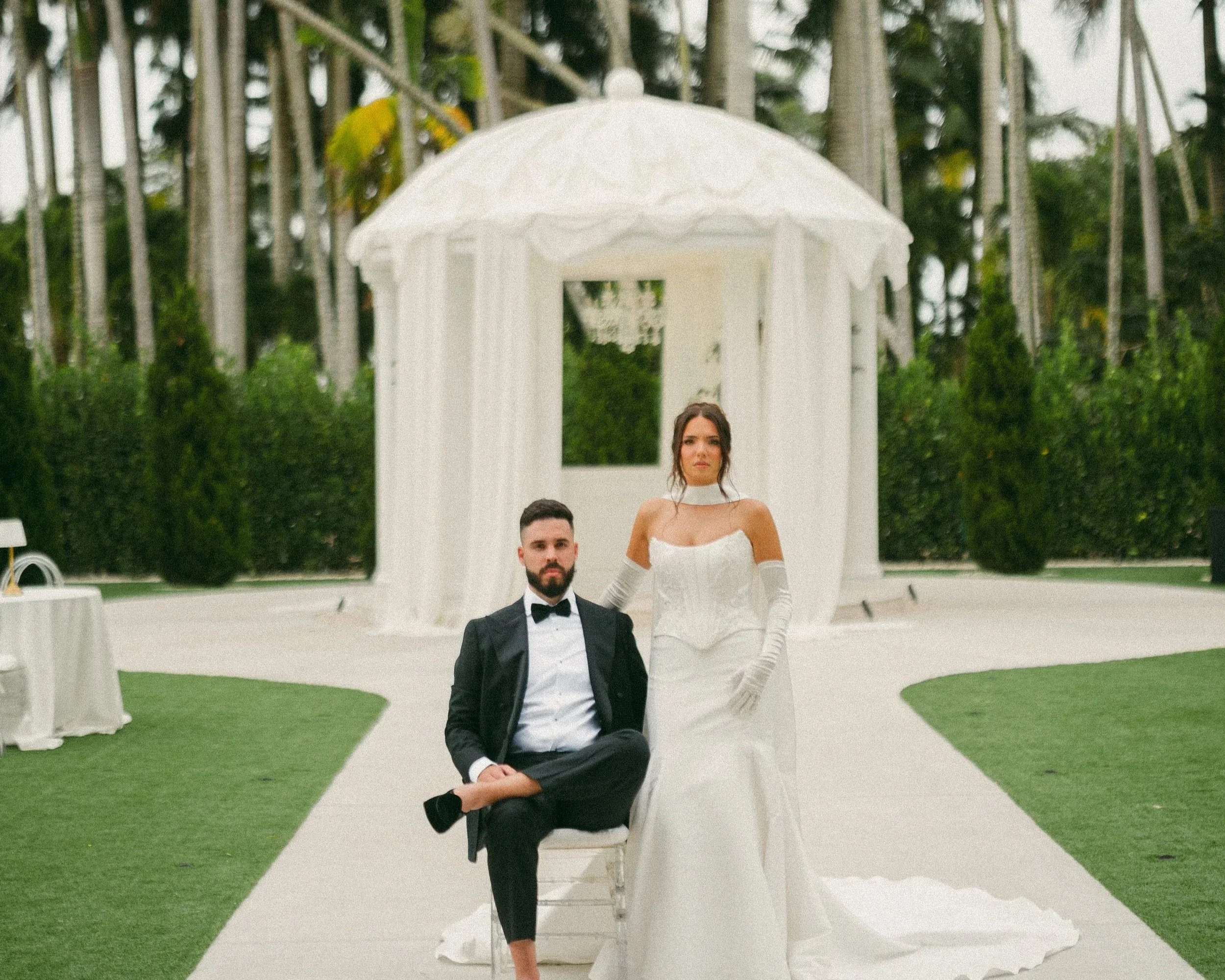 A bride and groom walking down the aisle at an outdoor wedding ceremony, surrounded by guests and palm trees.