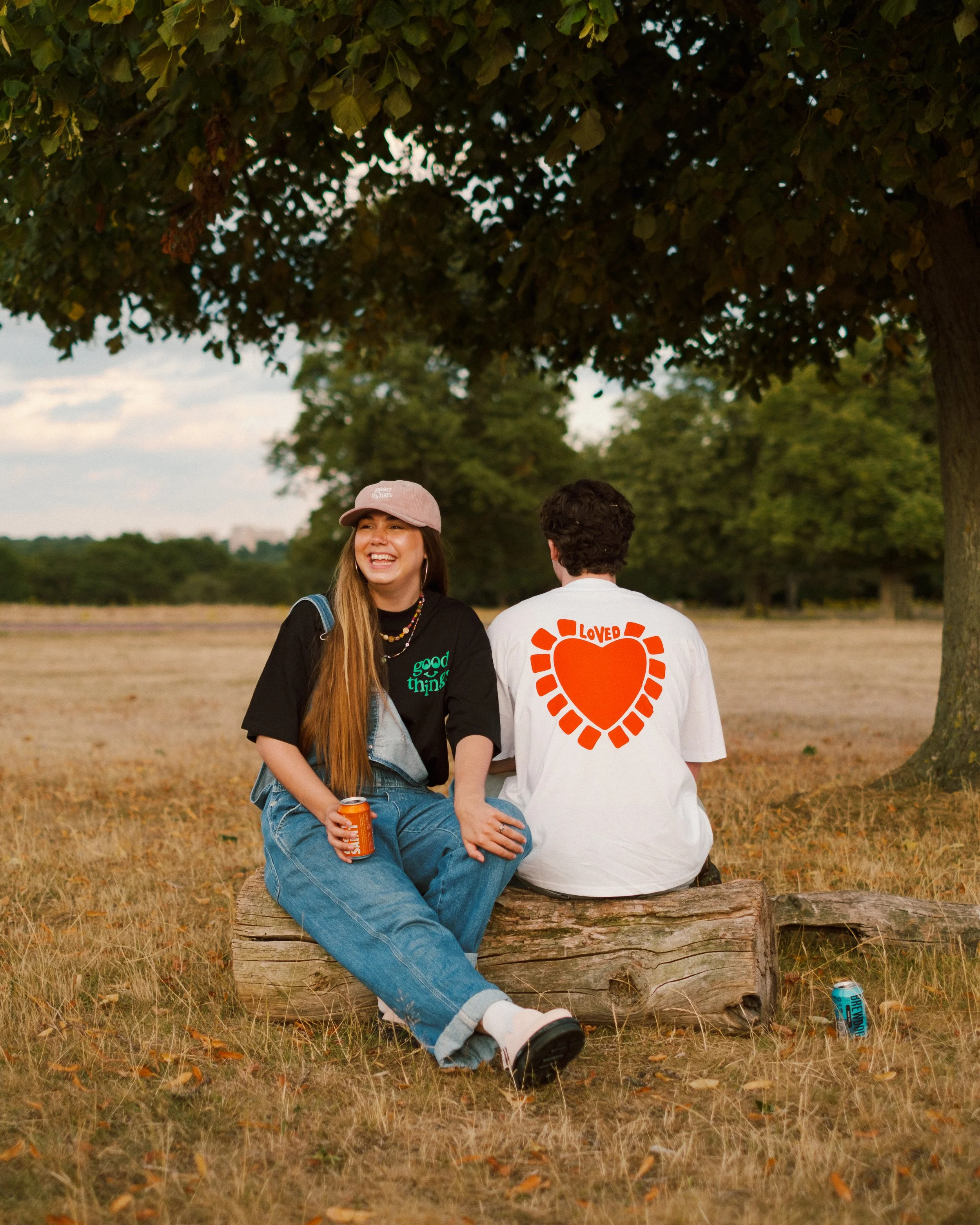 A smiling woman with long hair, wearing a pink baseball cap, black t-shirt, and jeans, sitting on a wooden log with a man whose back faces the camera. The man is wearing a white t-shirt with a large orange heart and the word 'LOVED' on the back, seat