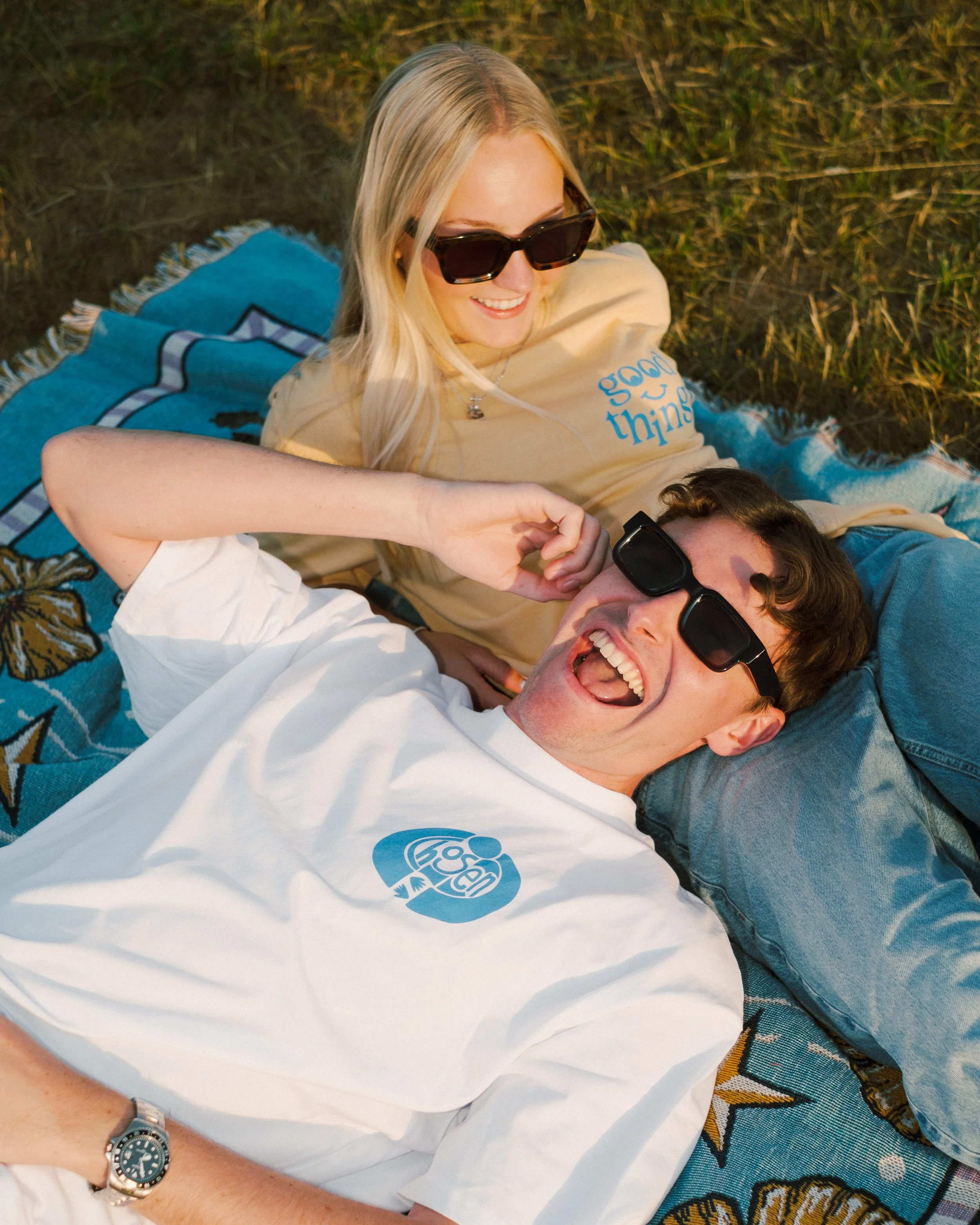 A young man and woman relaxing outdoors on a blanket, both wearing sunglasses and laughing.