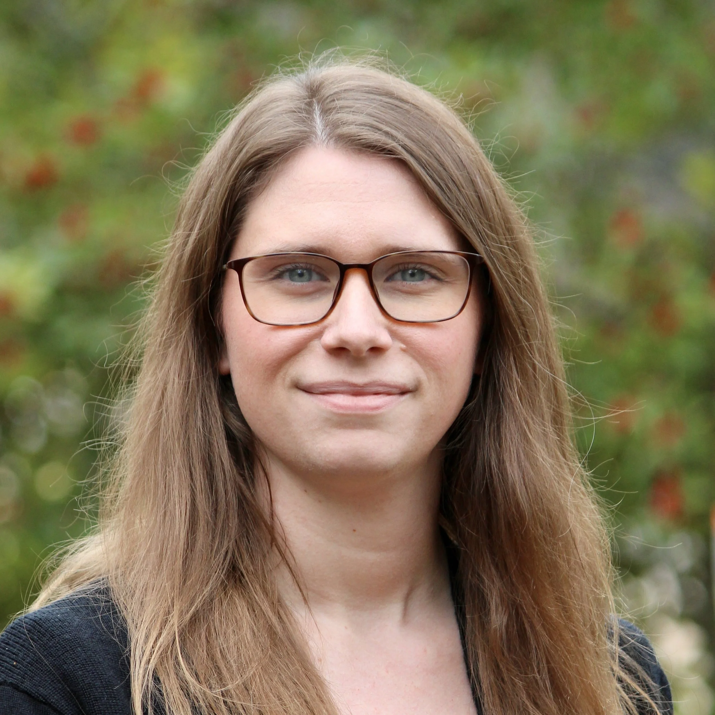 Portrait of a woman with long light brown hair, glasses, wearing a black top, outdoors with blurred green and red foliage in the background.