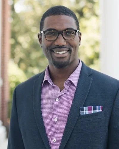 Walter Strickland with glasses, a beard, and short hair smiling outdoors, wearing a dark blazer over a pink shirt with a pocket square.