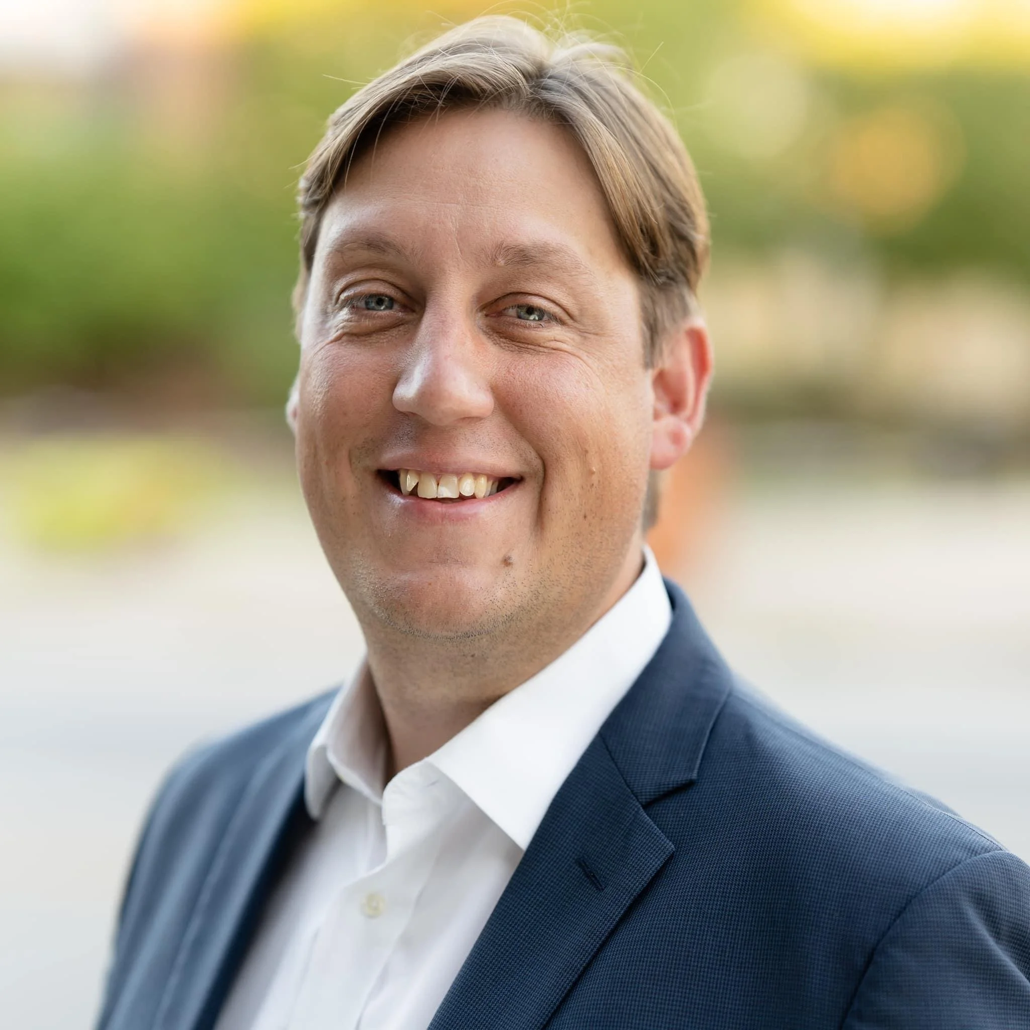 Close-up of Mark Caitlin with light brown hair in a suit and white shirt, smiling outdoors with a blurred green background.