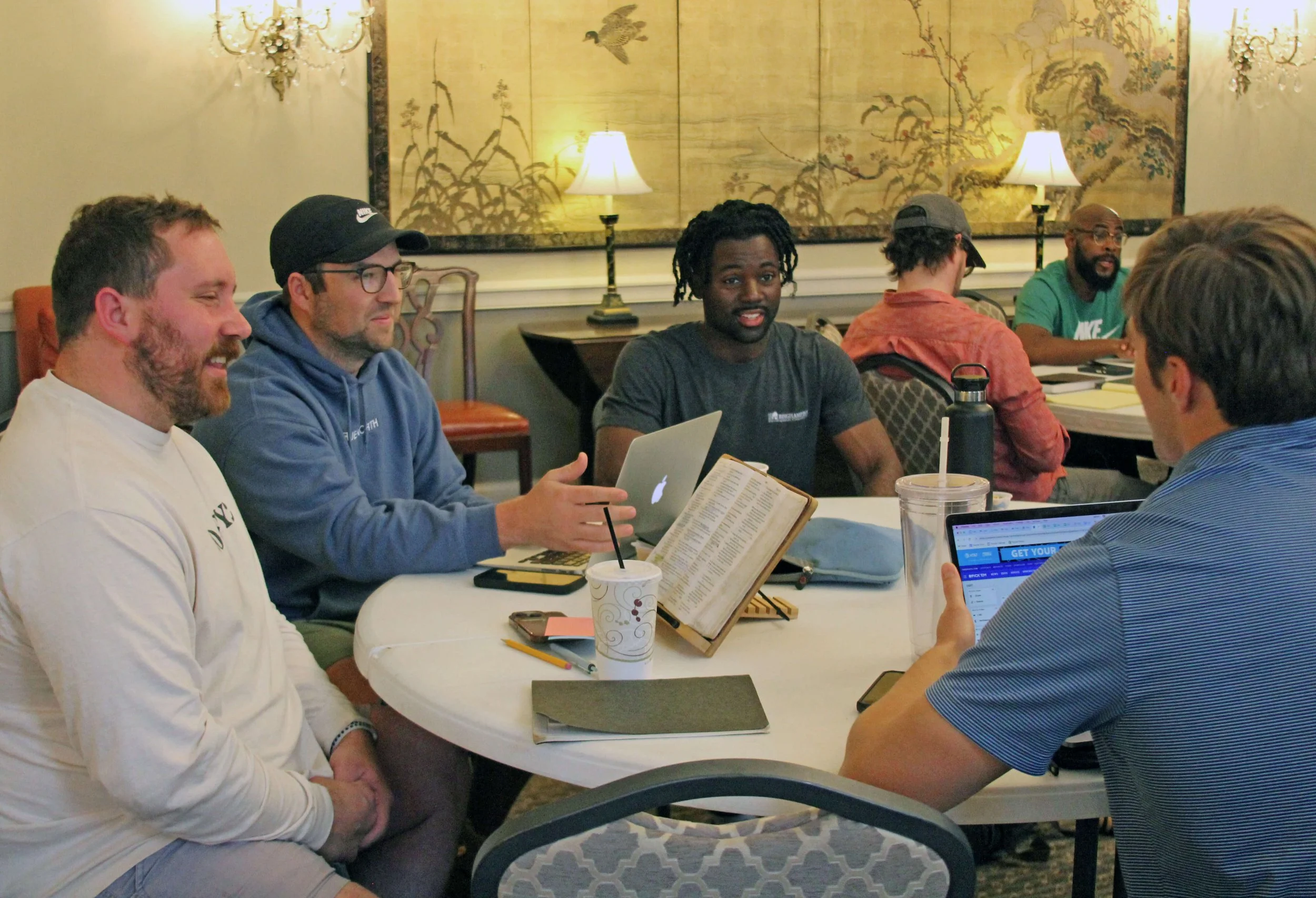 A group of six men sitting around a large white table engaged in discussion, with laptops, notebooks, and drinks on the table, in a room with elegant chandelier lighting and traditional artwork on the wall.