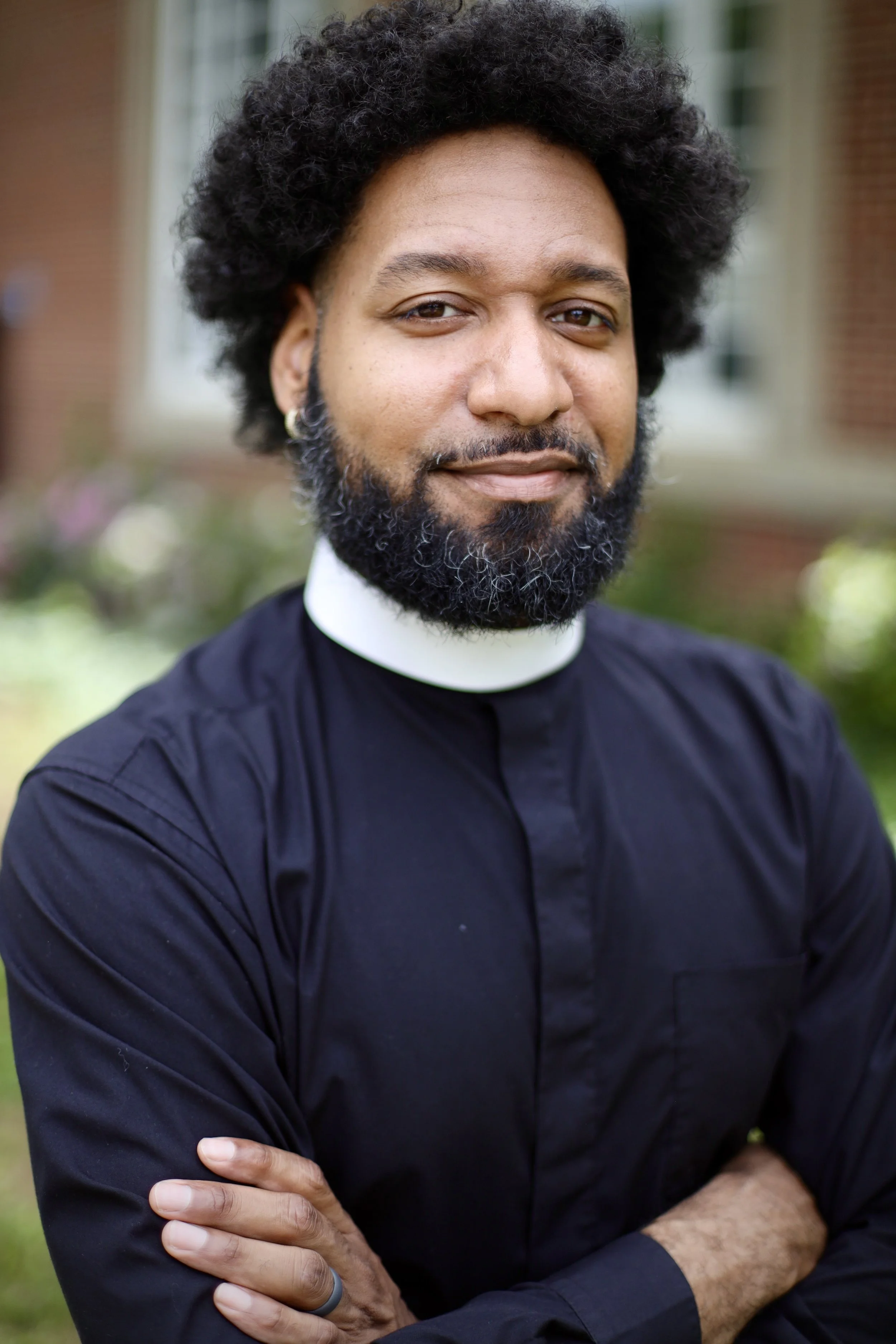 Close-up of Claude Atcho with a beard, wearing a clerical collar and a black shirt, crossing his arms outdoors.