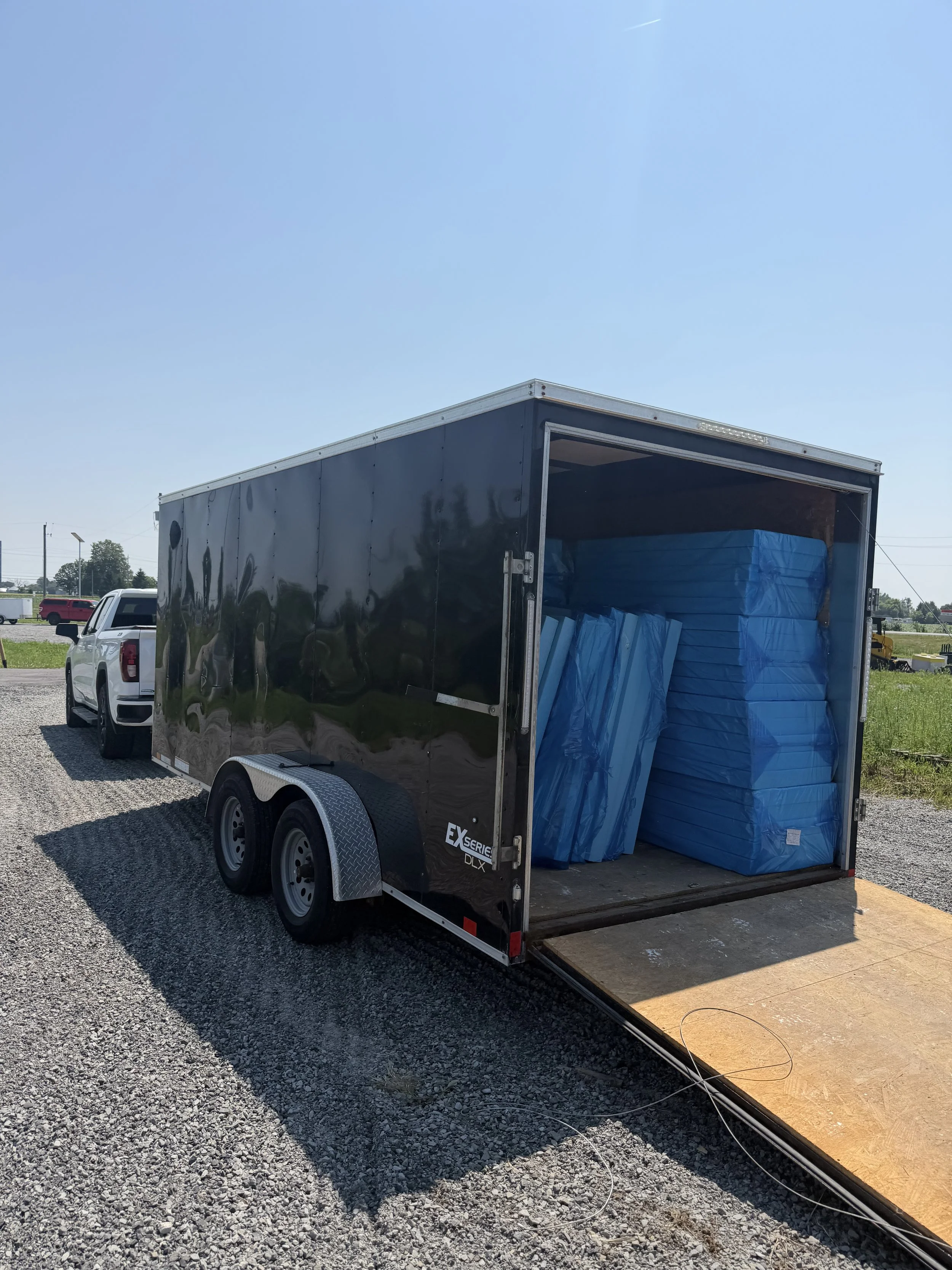 A black enclosed trailer with its rear door open, parked on a gravel lot, with blue packages or bins inside.