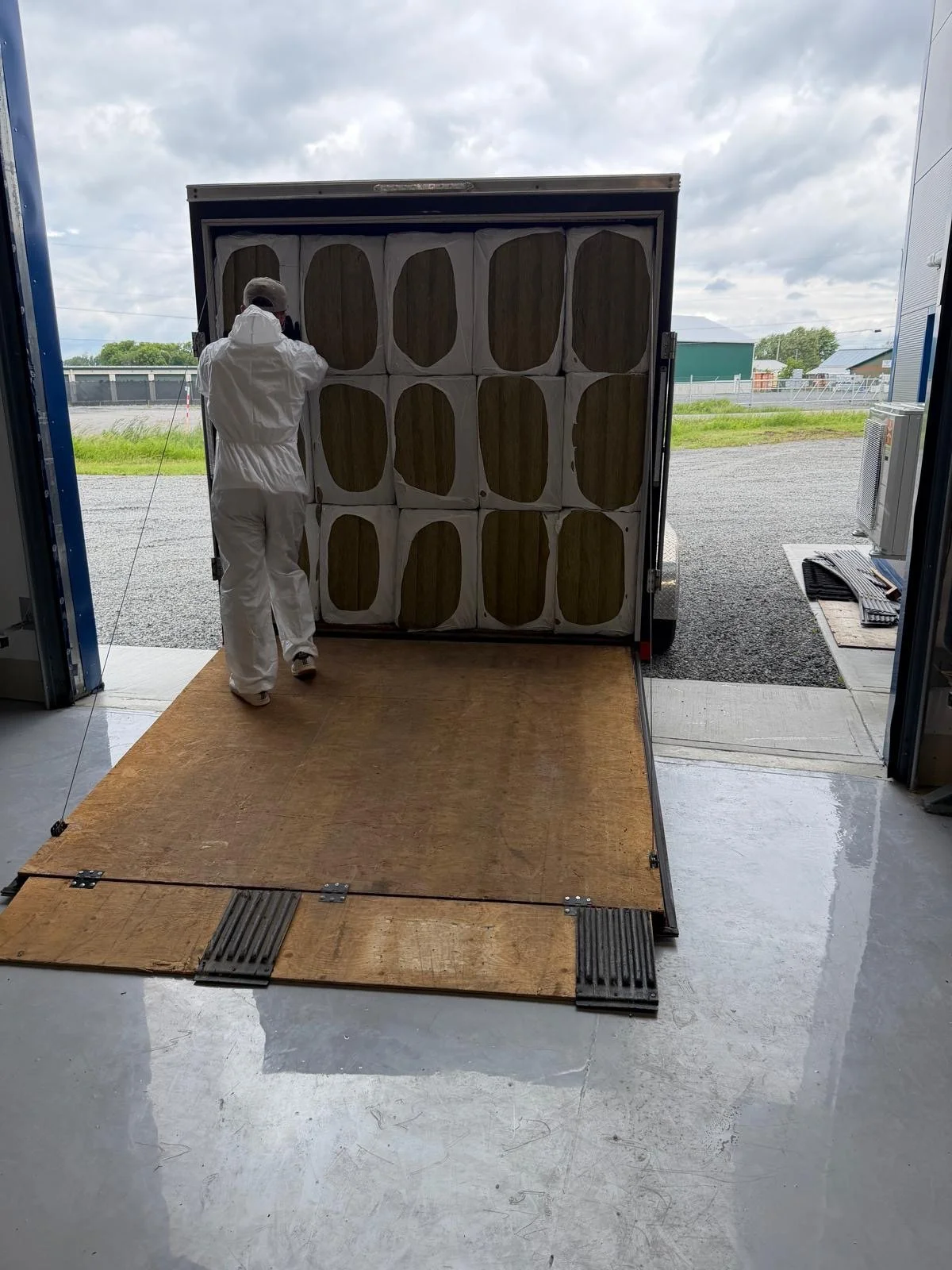 A person dressed in white protective clothing loading a trailer with large boxes that appear to contain insulation. The trailer is parked inside a building, with an open garage door revealing a cloudy sky and a rural landscape outside.