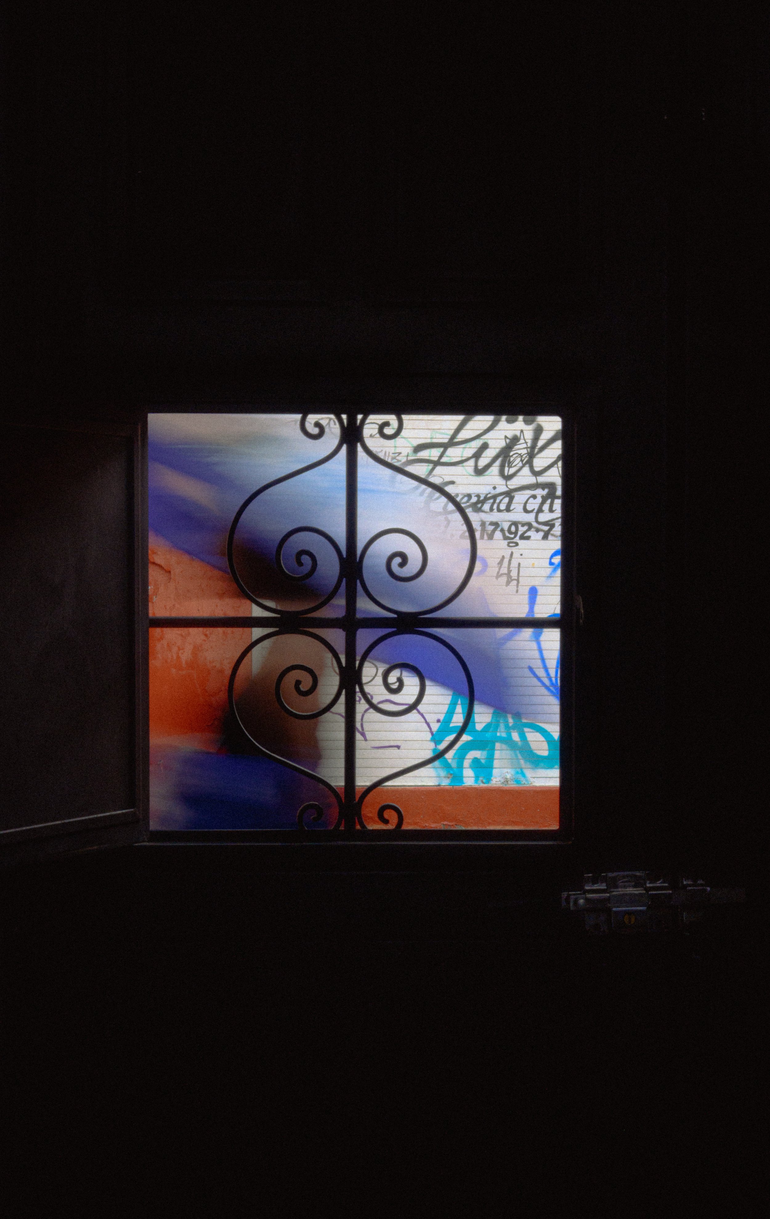  Vista de una ventana con reja de hierro decorativa, que muestra graffiti y letras manuscritas en una pared exterior.