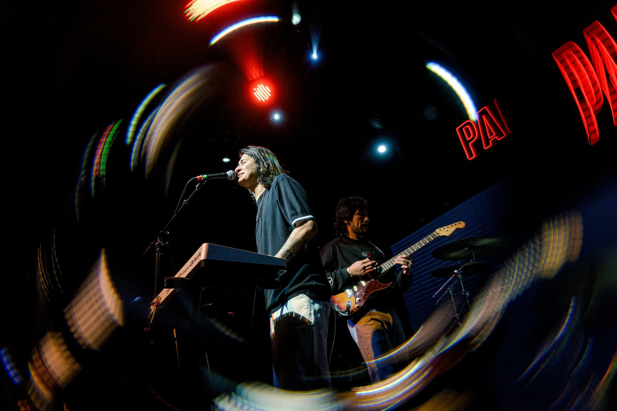 Dos músicos en un escenario, uno tocando teclado y cantando, el otro tocando guitarra eléctrica, con luces y textos neon rojos en el fondo.