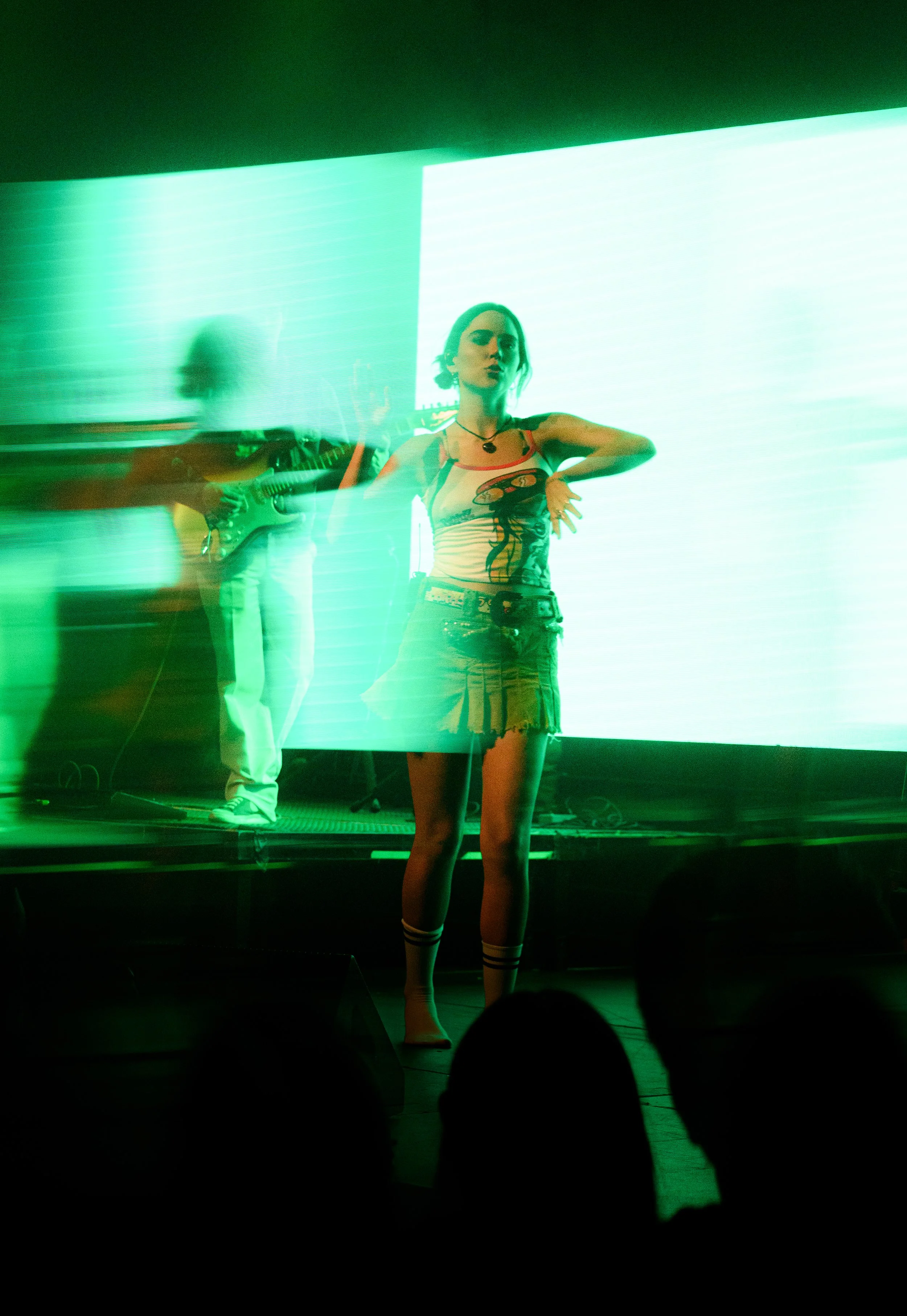 Una mujer en un escenario de concierto con un fondo de pantalla de luz verde, un hombre que toca el violín detrás y un público en primer plano.