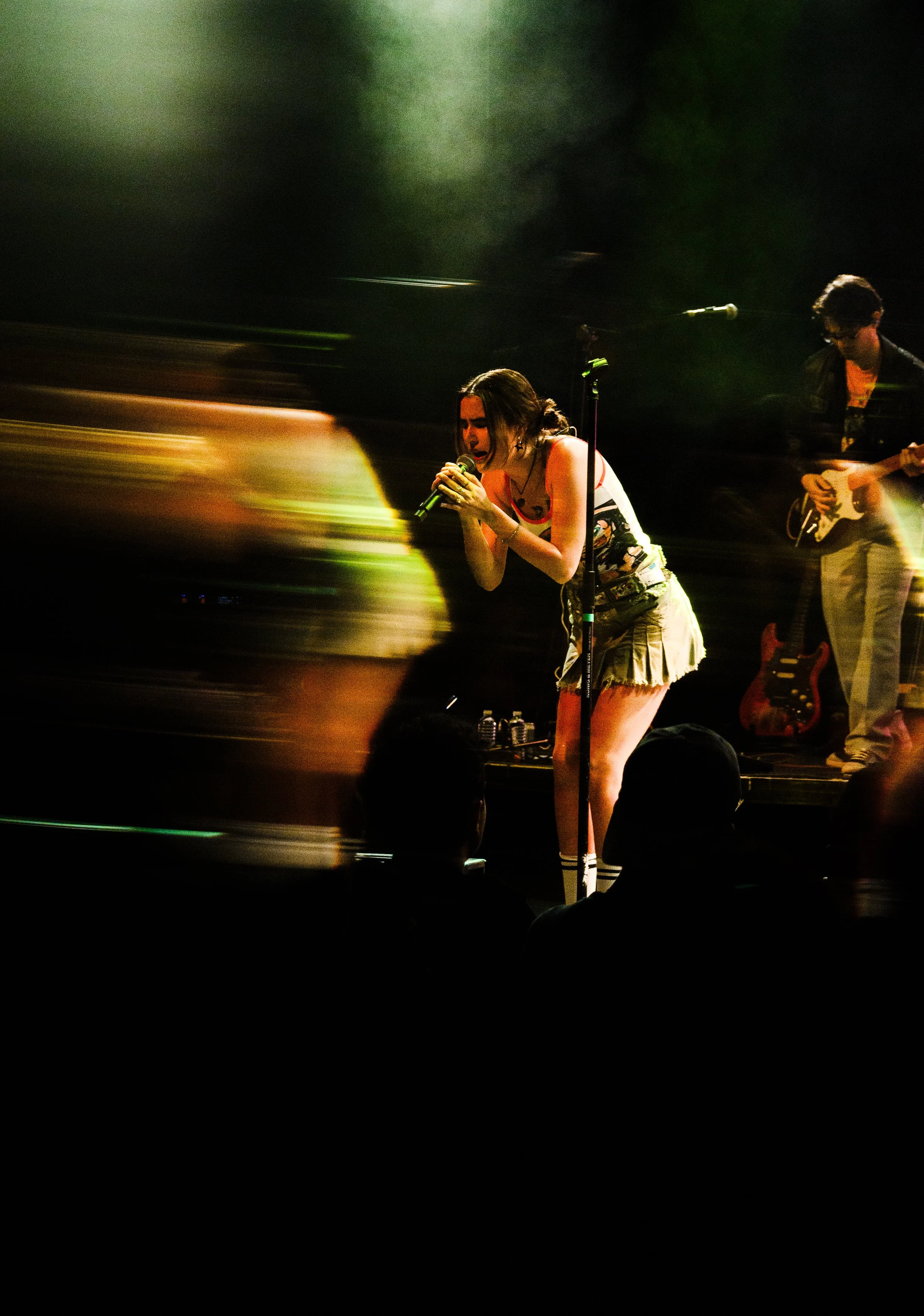 Una mujer canta en el escenario mientras toca la guitarra eléctrica, acompañada por un músico en el fondo. El escenario tiene luces y se observa la silueta de los espectadores.