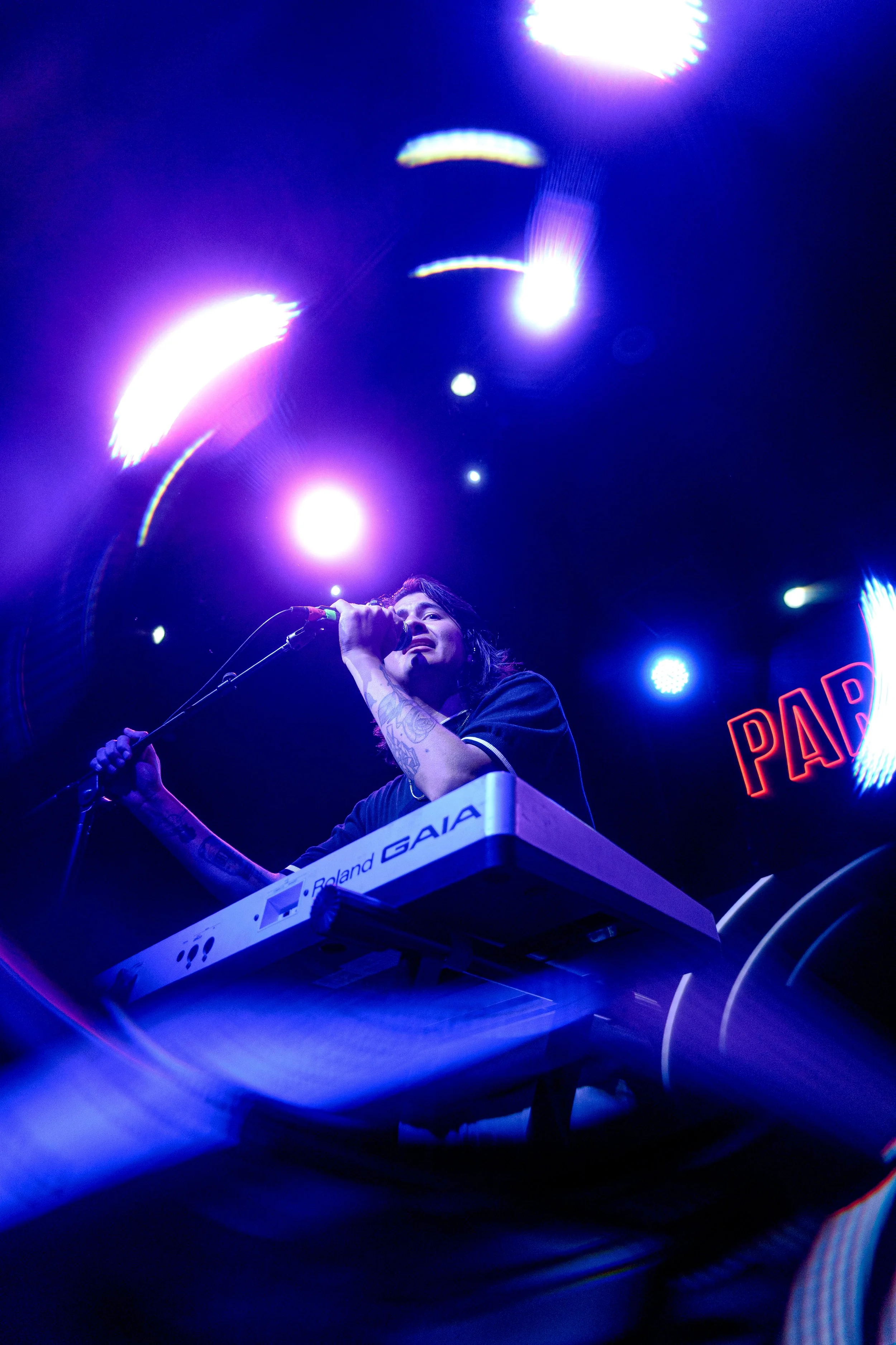 Un artista tocando un teclado en un escenario con luces brillantes y un cartel que dice 'PAP' en el fondo.